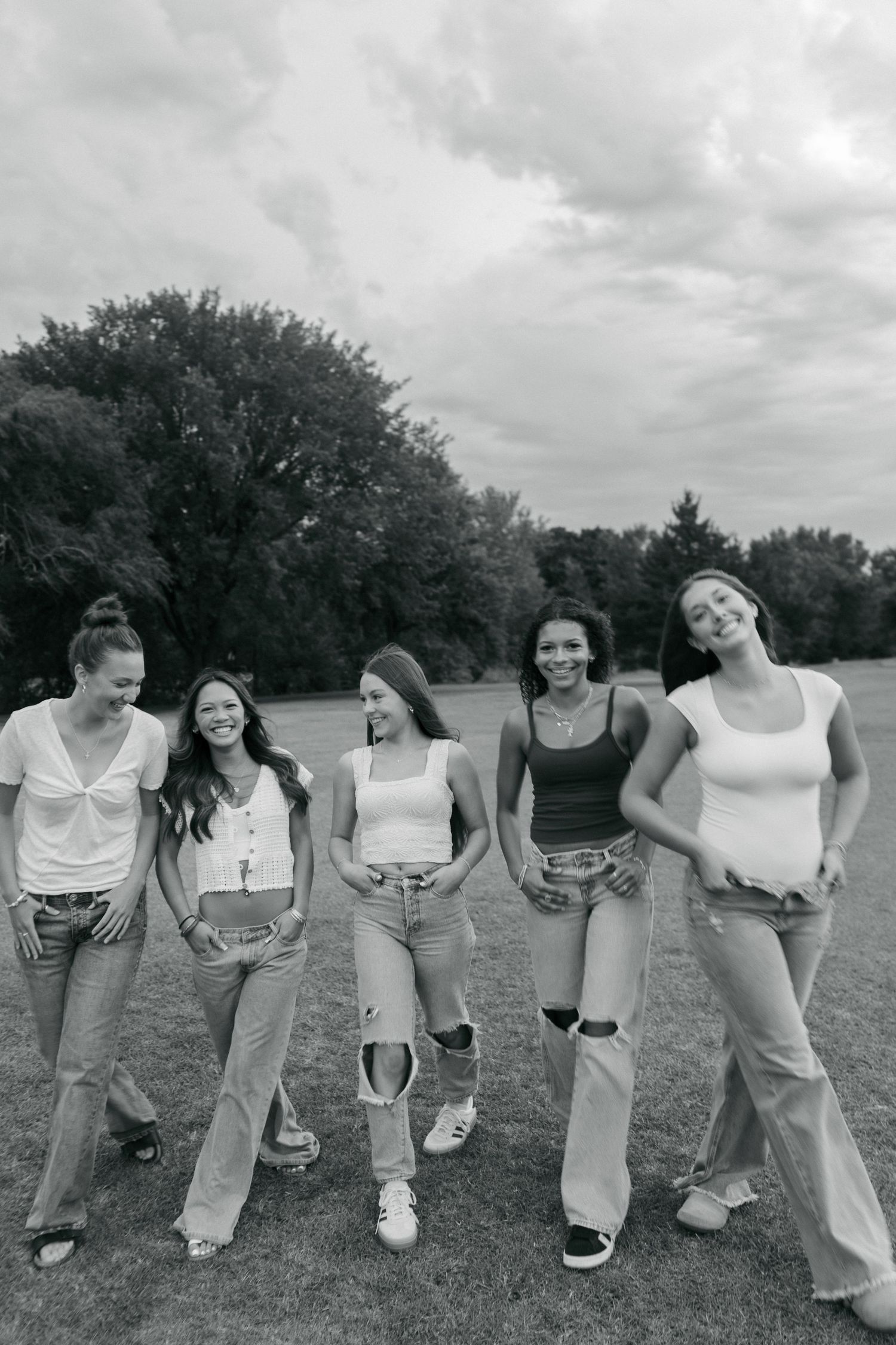 Five smiling young women posing together in a park wearing jeans and casual tops in black and white.