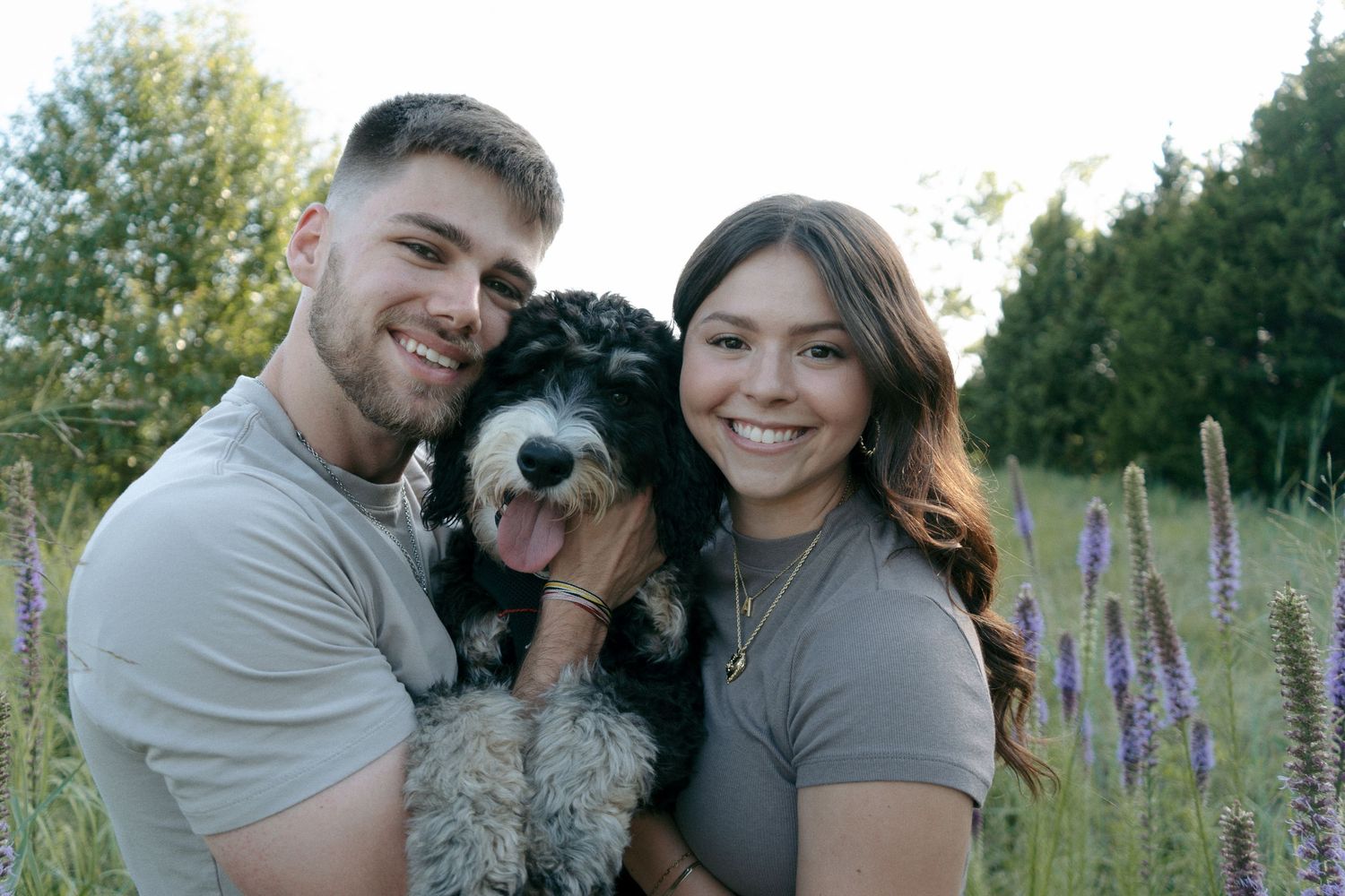 Smiling couple in matching gray shirts holding a fluffy black and white Bernedoodle dog outdoors.