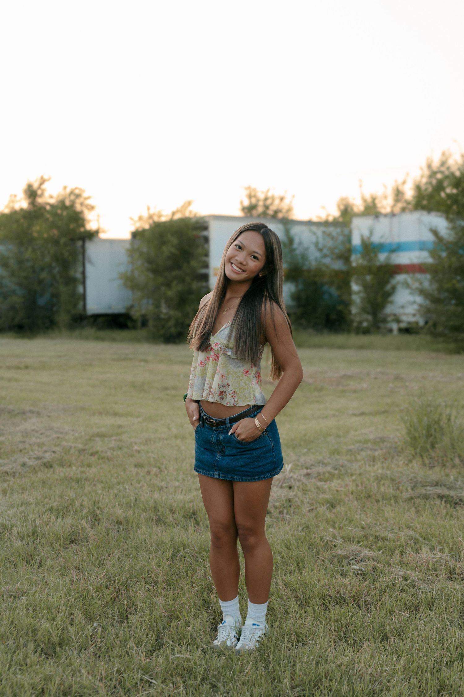 Young woman in floral crop top and denim skirt smiling in a grassy field at sunset.
