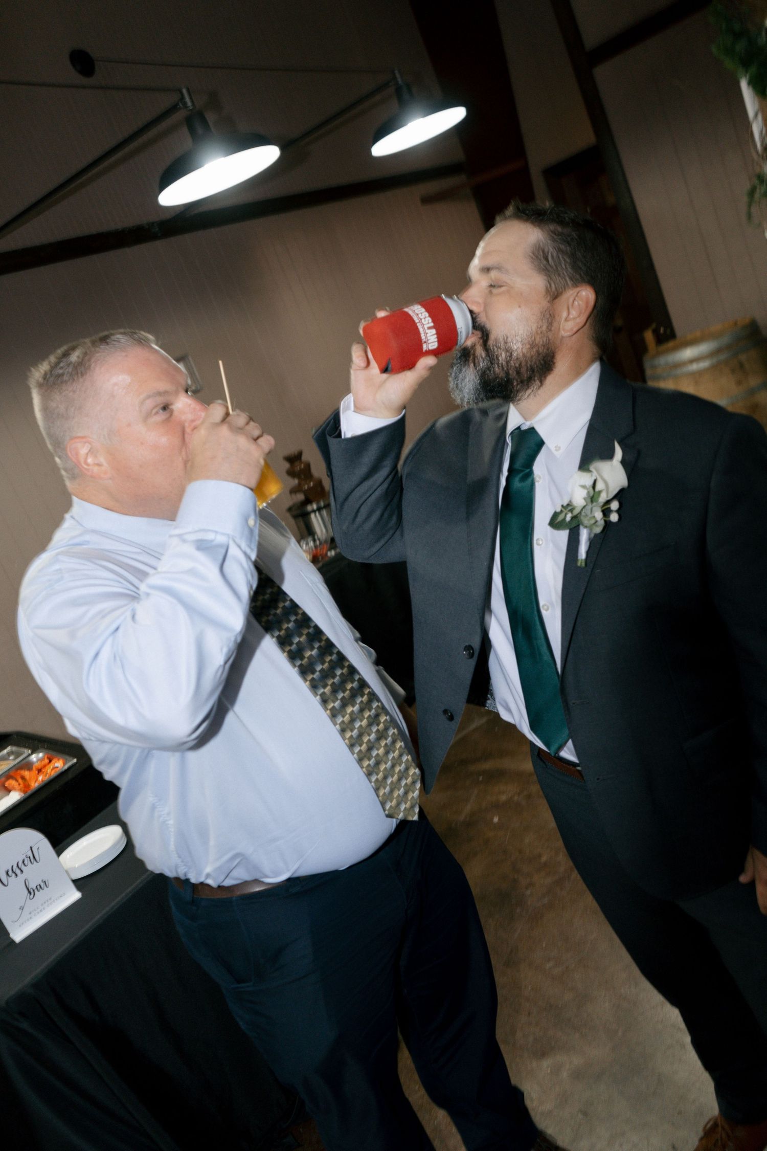 Two men in suits drinking from cups at a wedding reception, one wearing a boutonniere with a green tie.