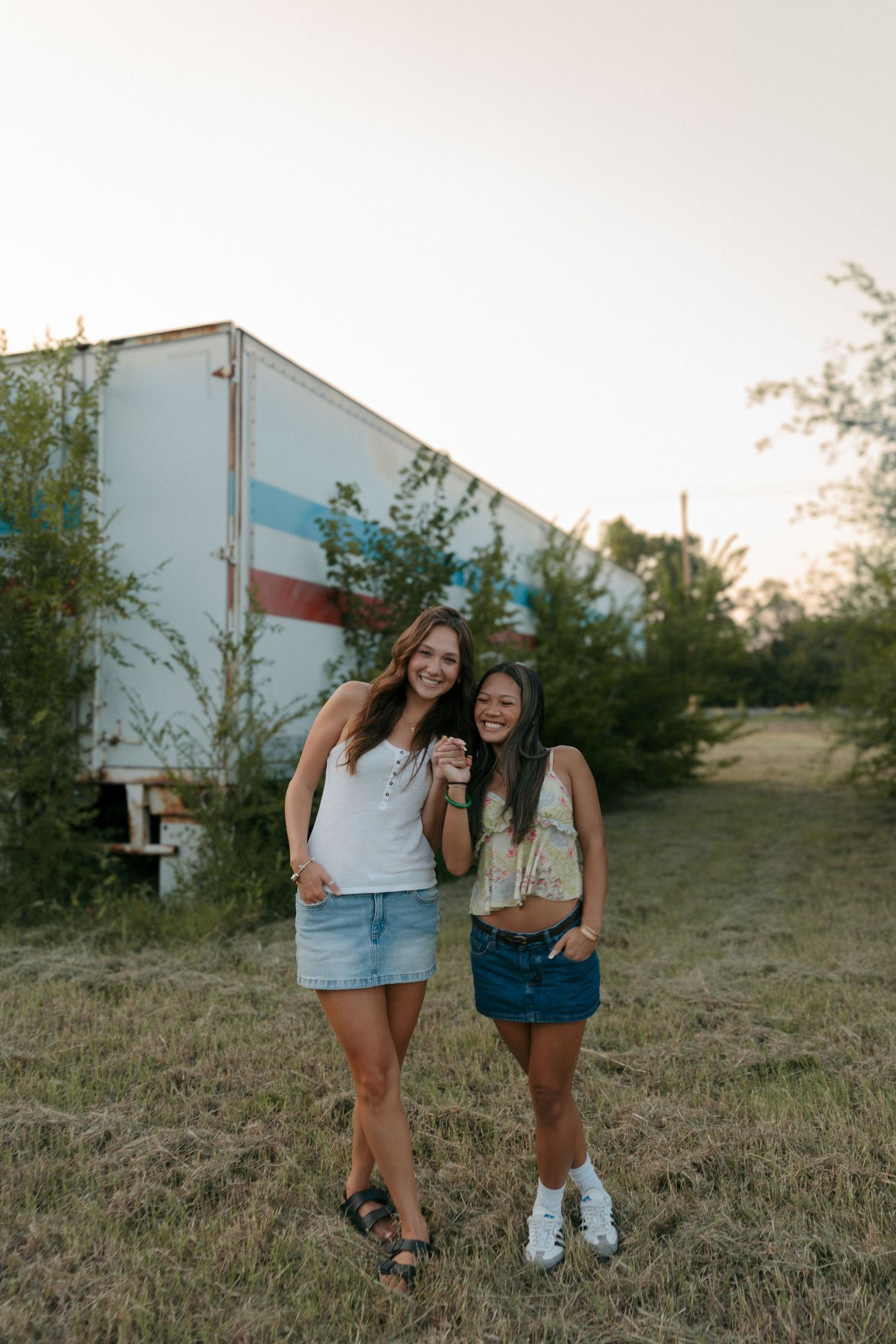 Two young women in denim skirts smiling outdoors near a trailer at sunset.
