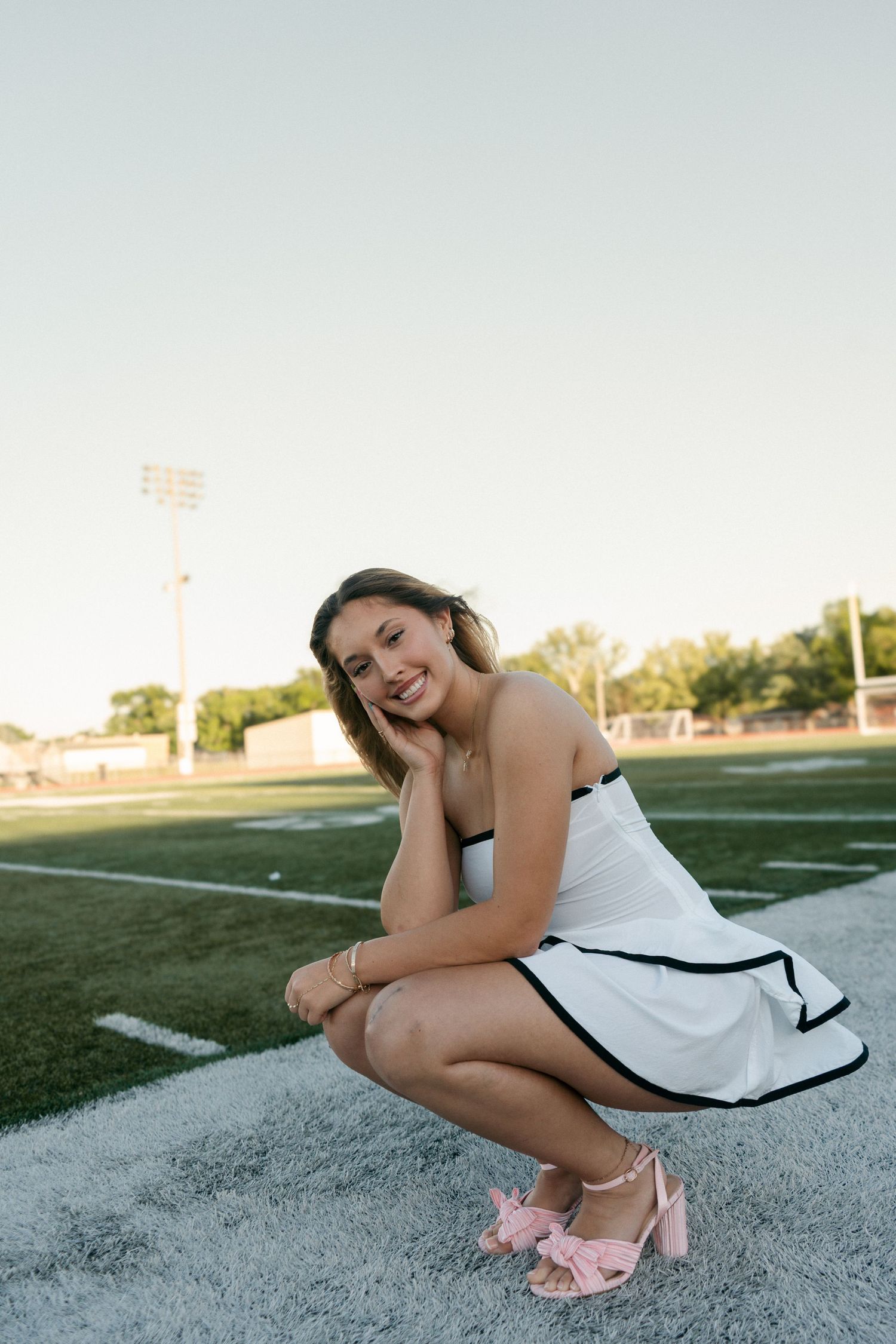 Young woman in white strapless dress and pink bow heels posing on football field sideline.