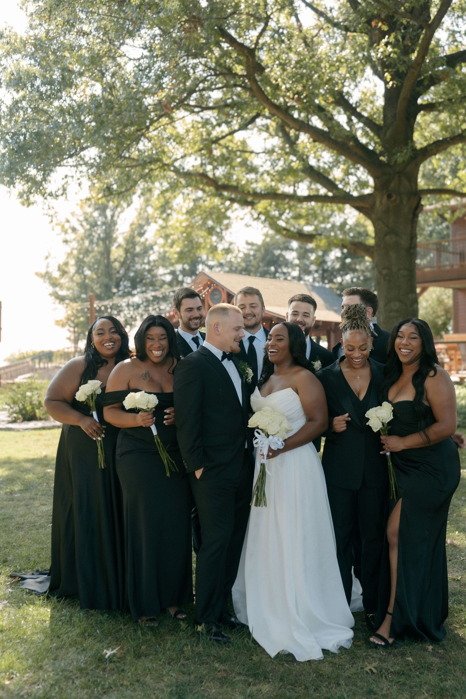 Wedding party in black attire with bride in white gown posing under oak tree at outdoor venue.