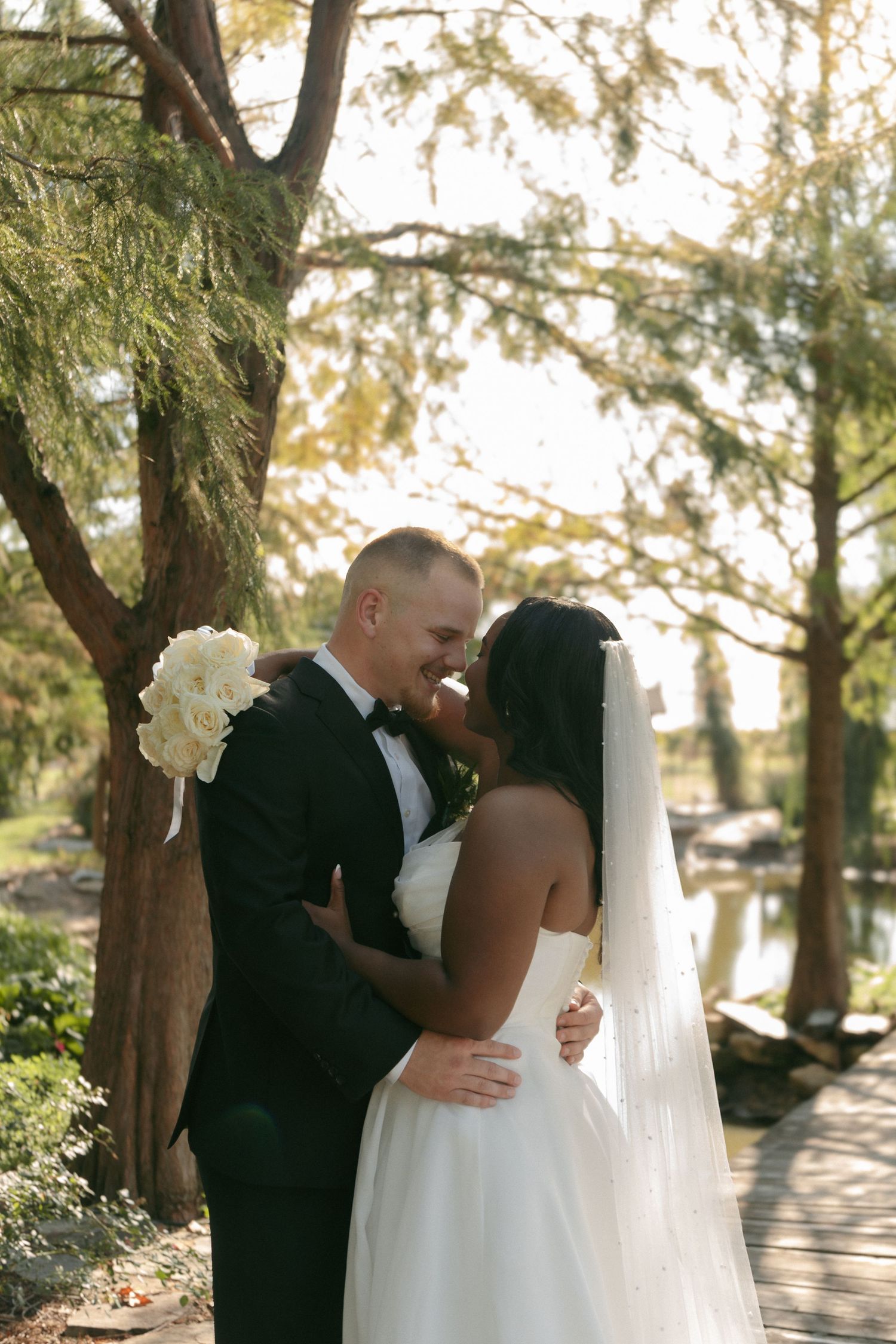 Interracial bride and groom sharing intimate moment nearly kissing outdoors near pond and trees.