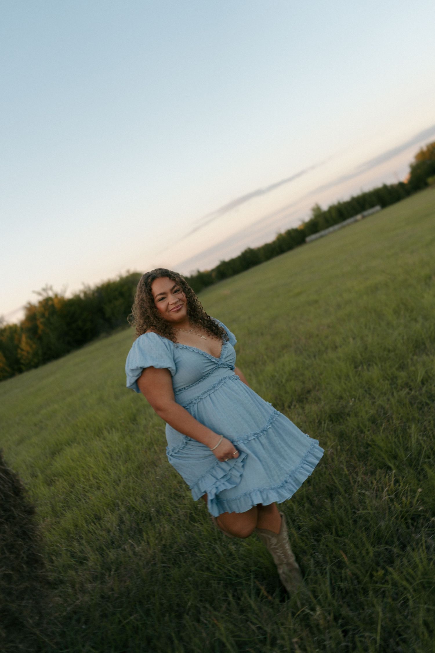 Young woman in light blue midi dress twirling in a green field near a hay bale at dusk.