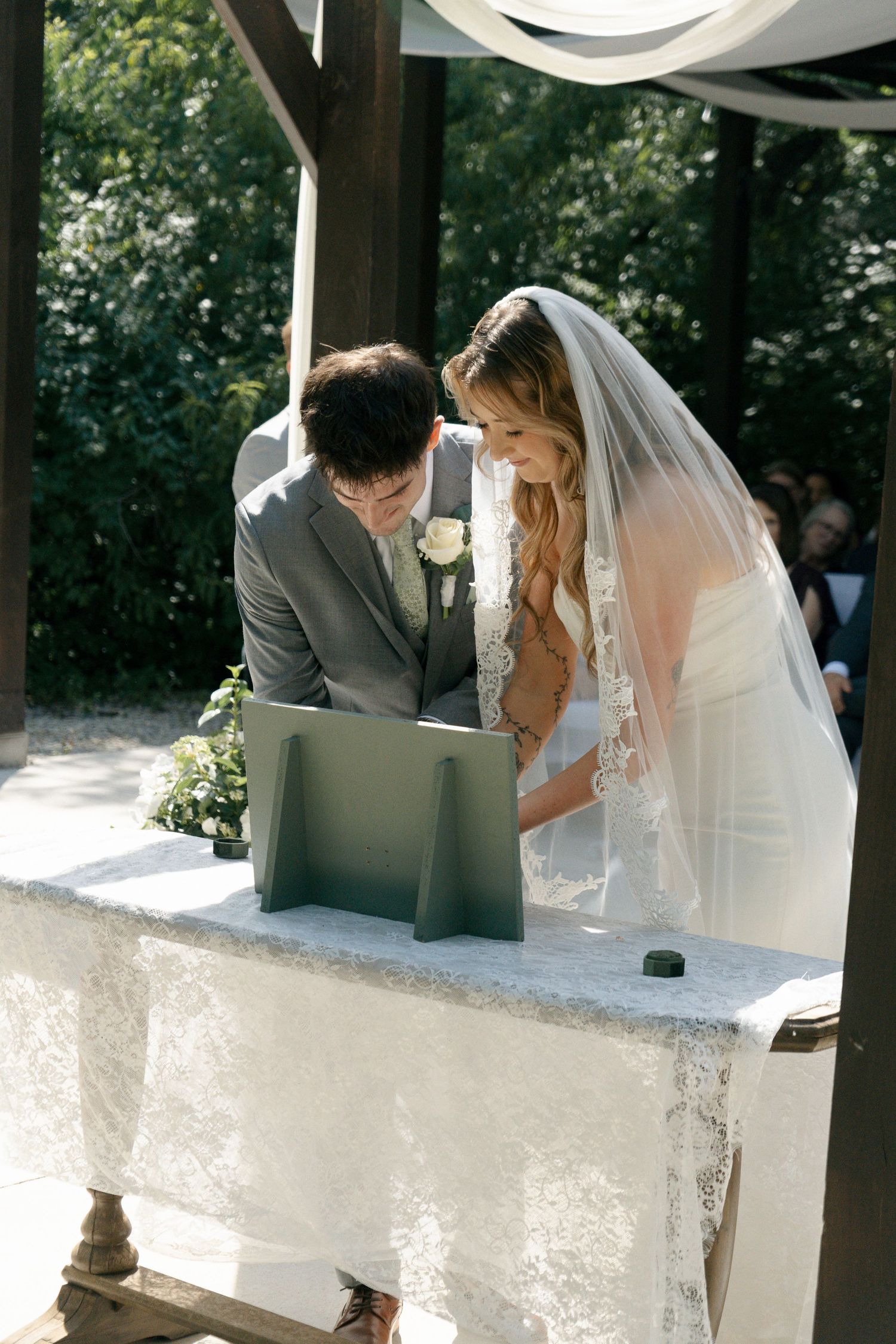 Bride and groom signing wedding register at outdoor ceremony altar, bride wearing lace veil and white gown.