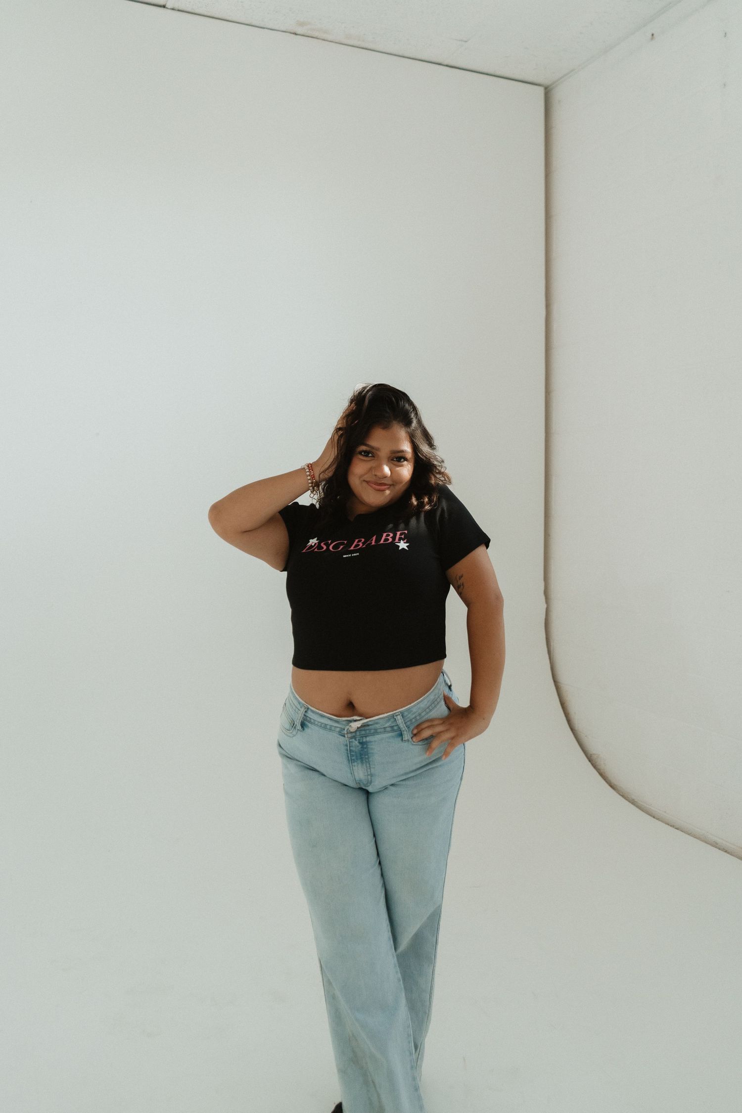 Young woman in black crop top and light blue jeans posing confidently in a white studio setting.