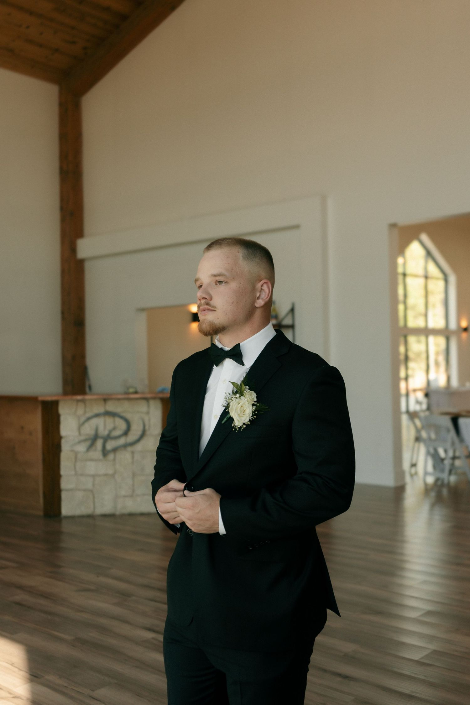 Groom in black tuxedo with boutonniere standing confidently inside elegant wedding venue.