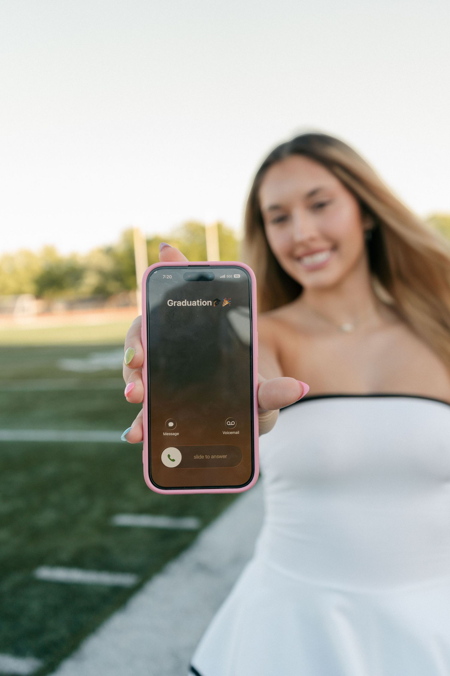 Young woman in white strapless dress holding pink iPhone displaying Graduation screen on sports field.