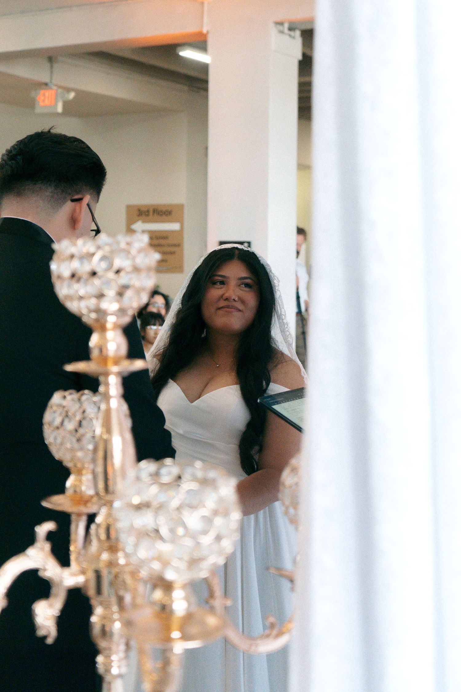 Bride in white off-shoulder gown exchanges vows with groom at elegant wedding ceremony with crystal candelabras.