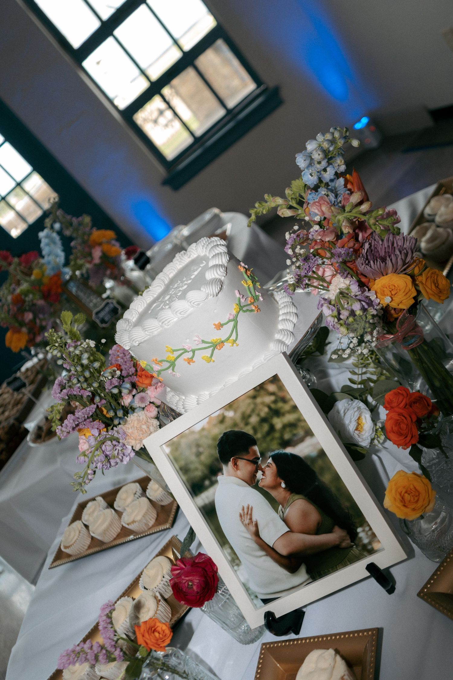 Elegant wedding cake table with colorful floral arrangements, cupcakes, and a romantic couple photo display at a reception.