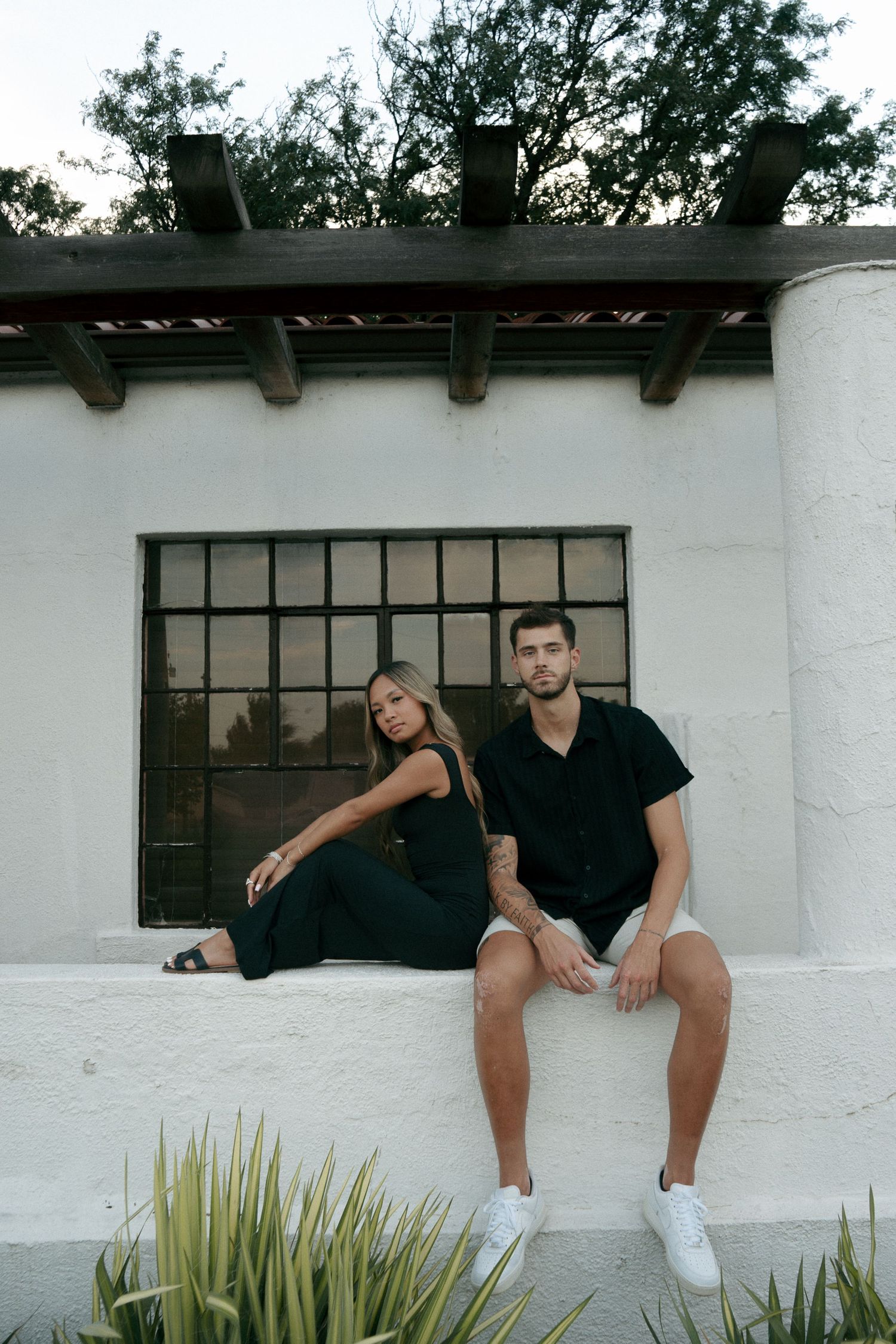 Couple in matching black outfits sitting on white ledge in front of industrial window outdoors.