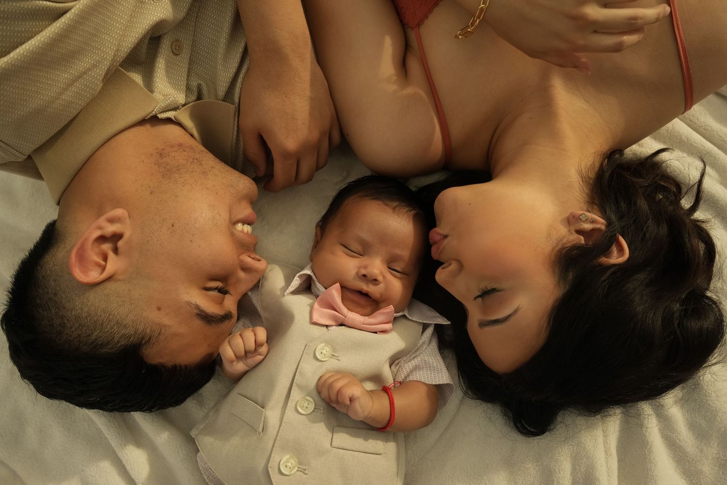 Happy parents lying with their smiling newborn baby on a white bed.