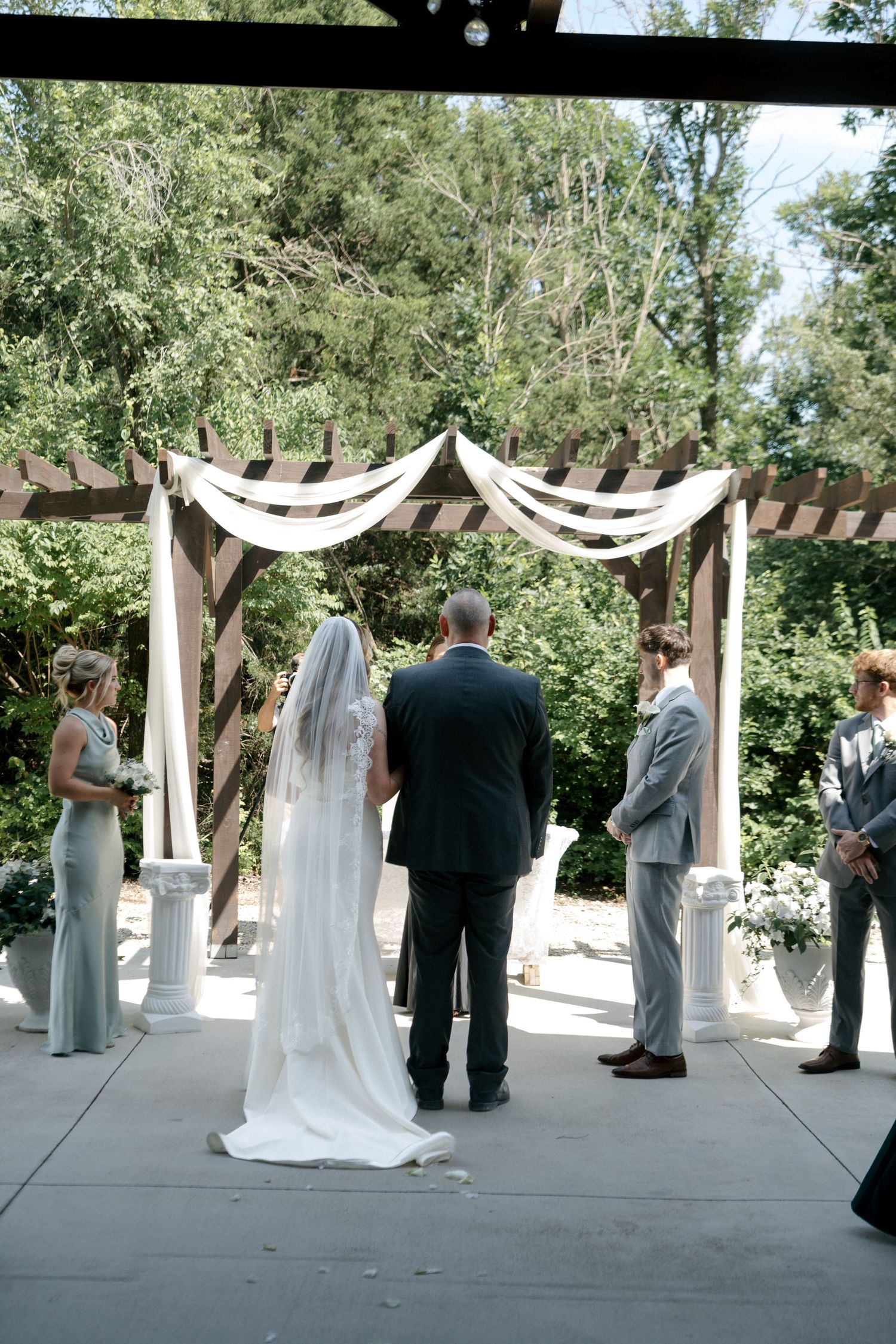 Bride and groom stand at outdoor wedding altar with draped pergola, flanked by bridesmaids and groomsmen amid lush greenery.