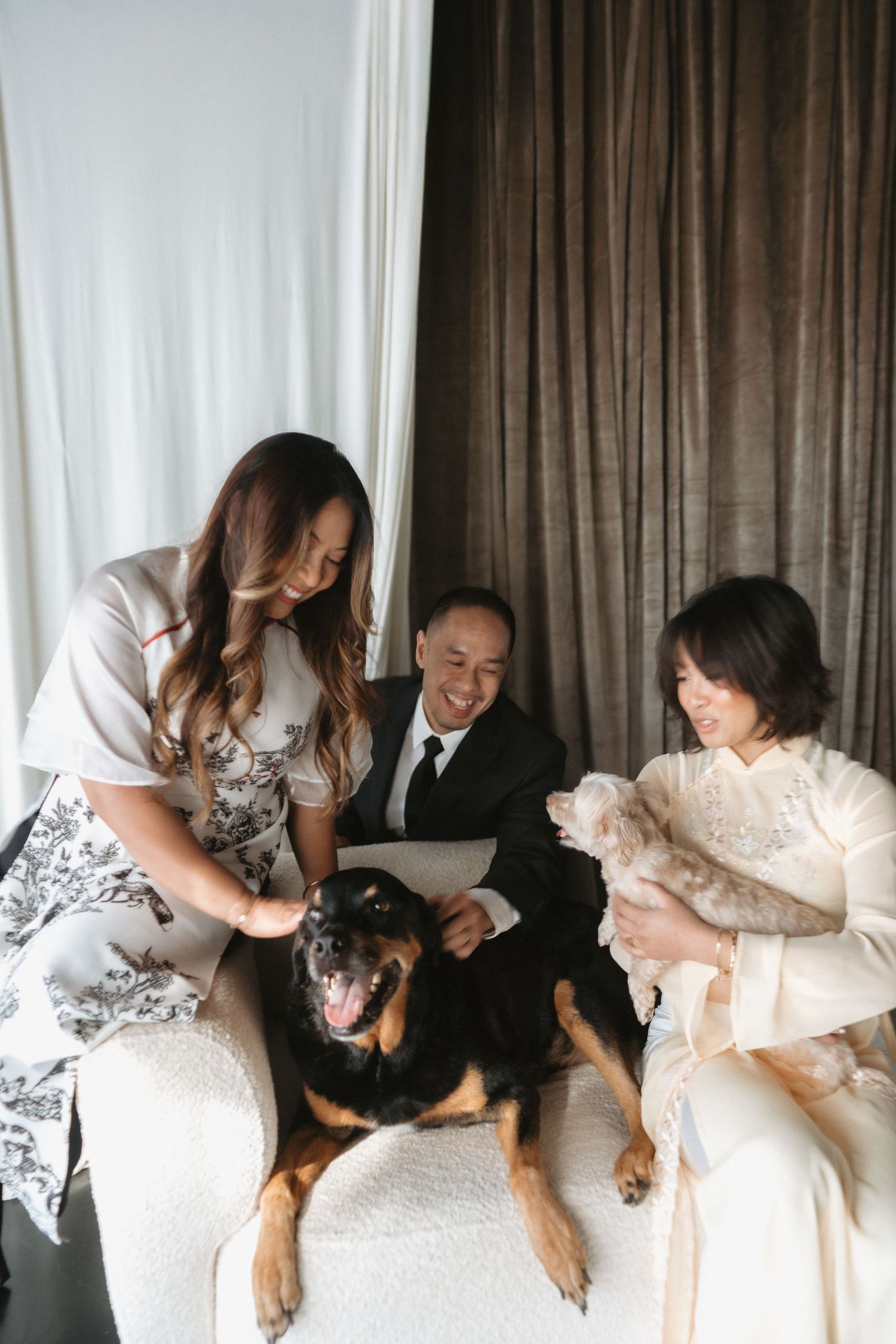 A bride, groom, and bridesmaid smile while petting a Rottweiler during a wedding photo session.