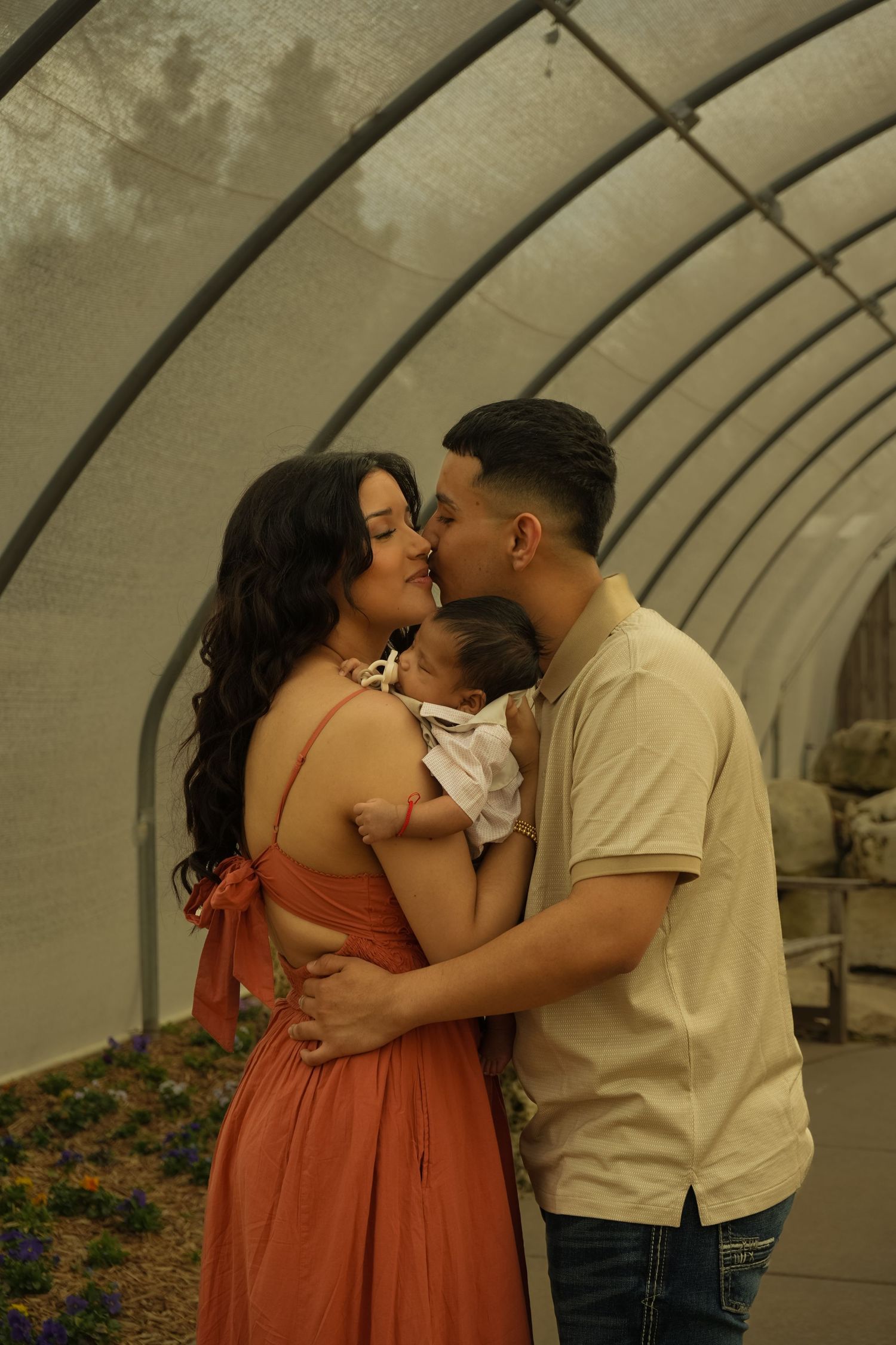 Couple sharing a kiss while holding newborn baby inside a greenhouse tunnel with flowers.