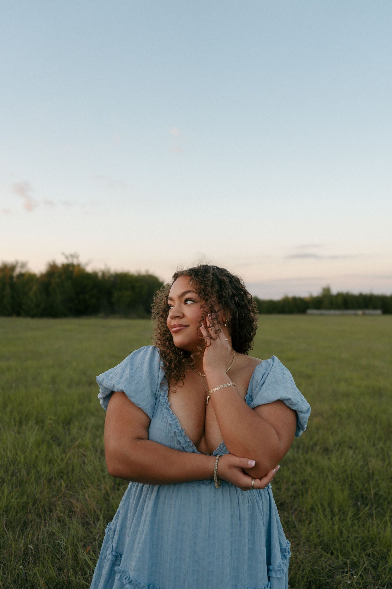 Curly haired woman in blue dress smiling in green field at sunset touching her face.