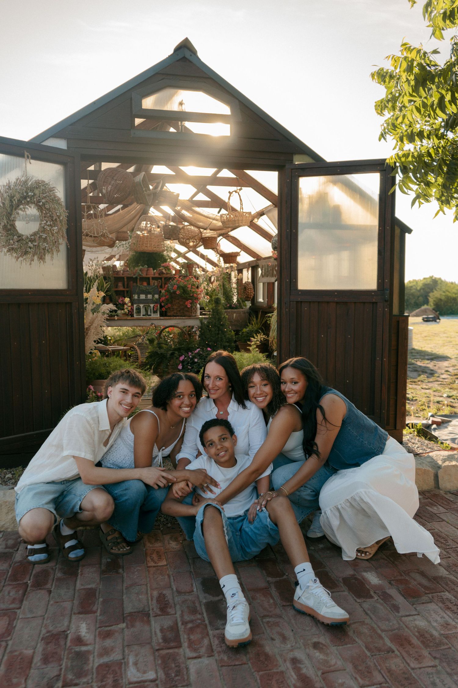 A joyful family group poses together in front of a rustic greenhouse, smiling warmly in golden hour sunlight.