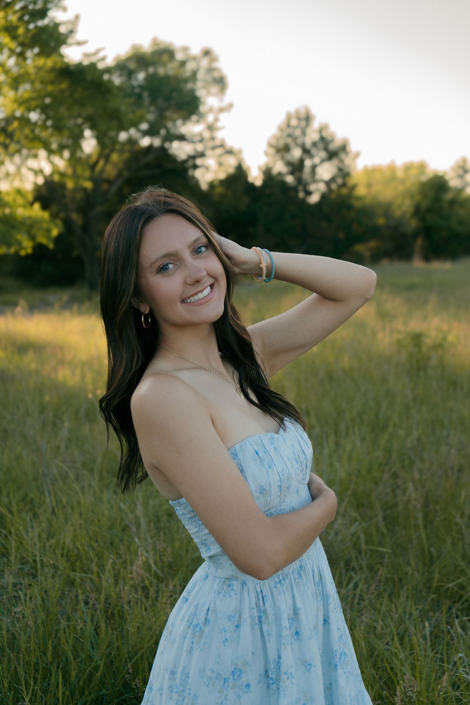 Smiling brunette woman in strapless blue floral dress posing in sunlit green meadow at dusk.