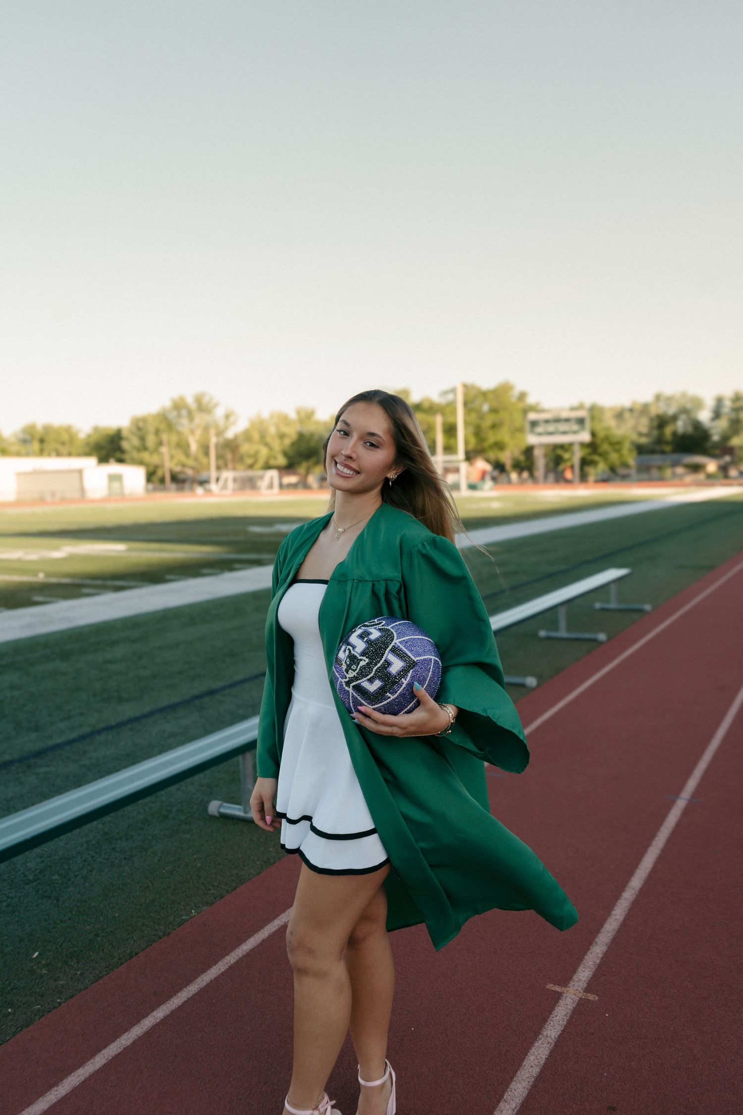 Smiling graduate in green cap and gown holds volleyball on school track.