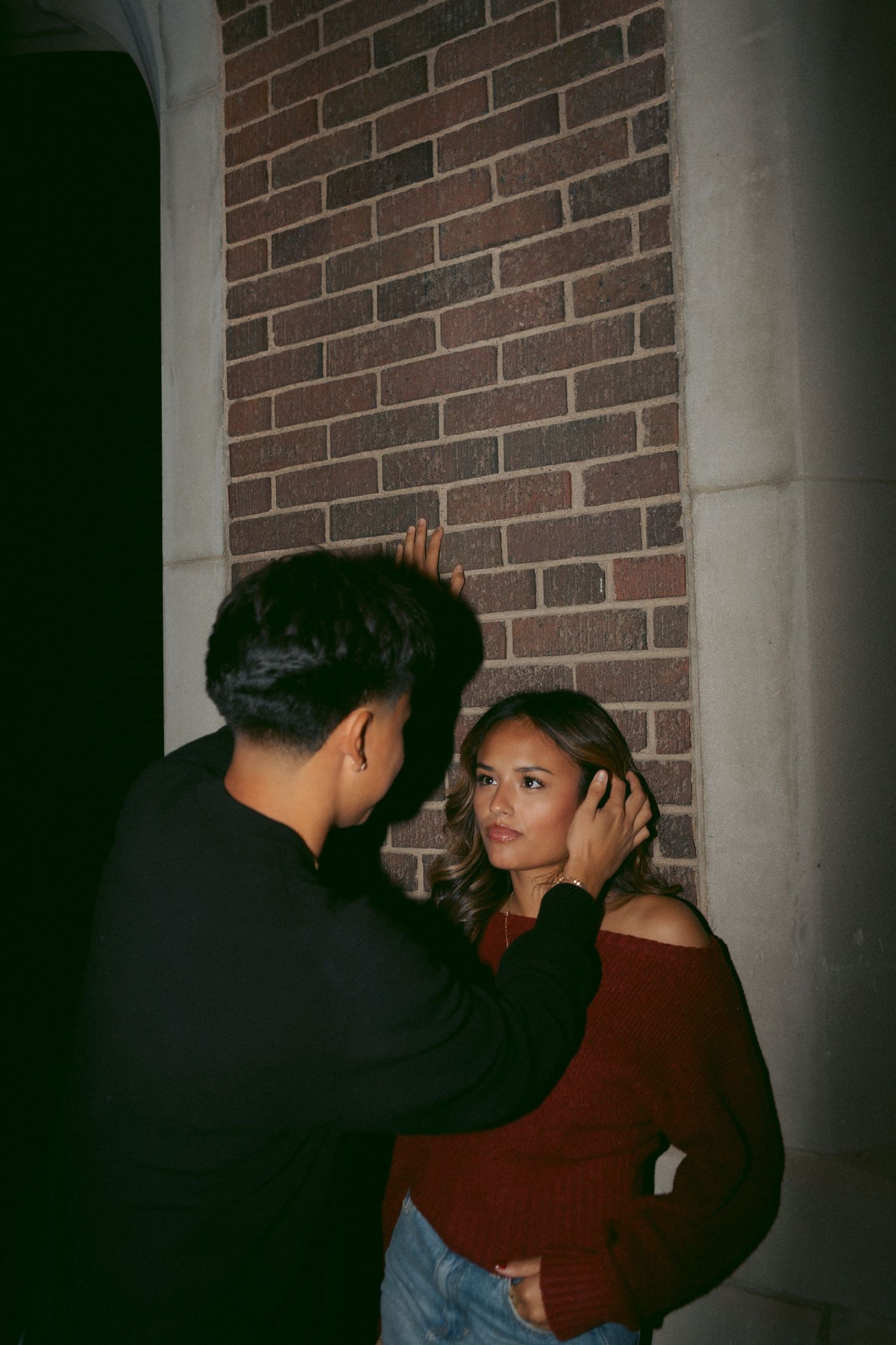 Intimate moment of a young man gently touching his girlfriends face against a brick wall at night.