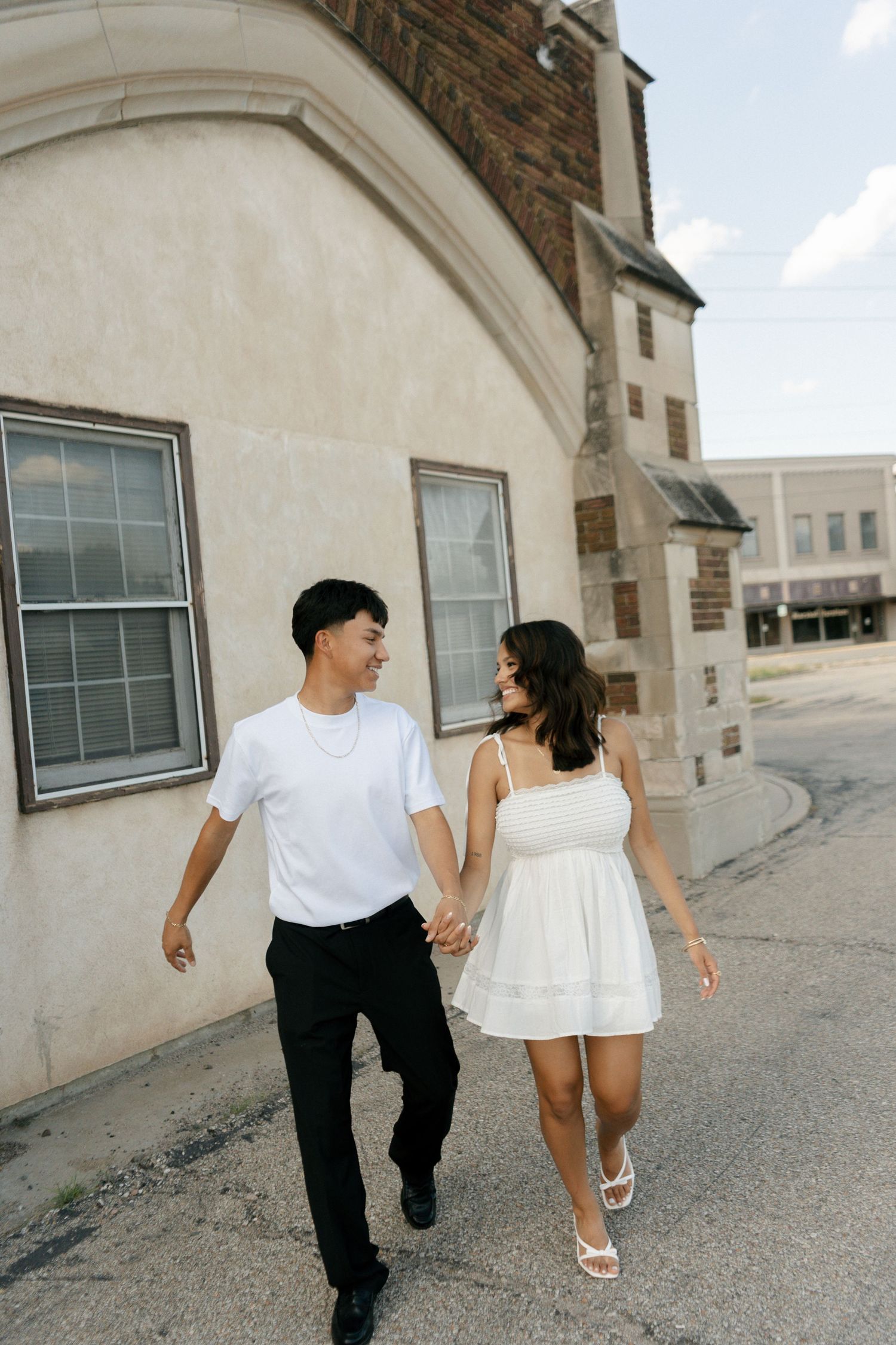 A couple holds hands and walks together near a building, the man in white tee and black pants, woman in white mini dress.