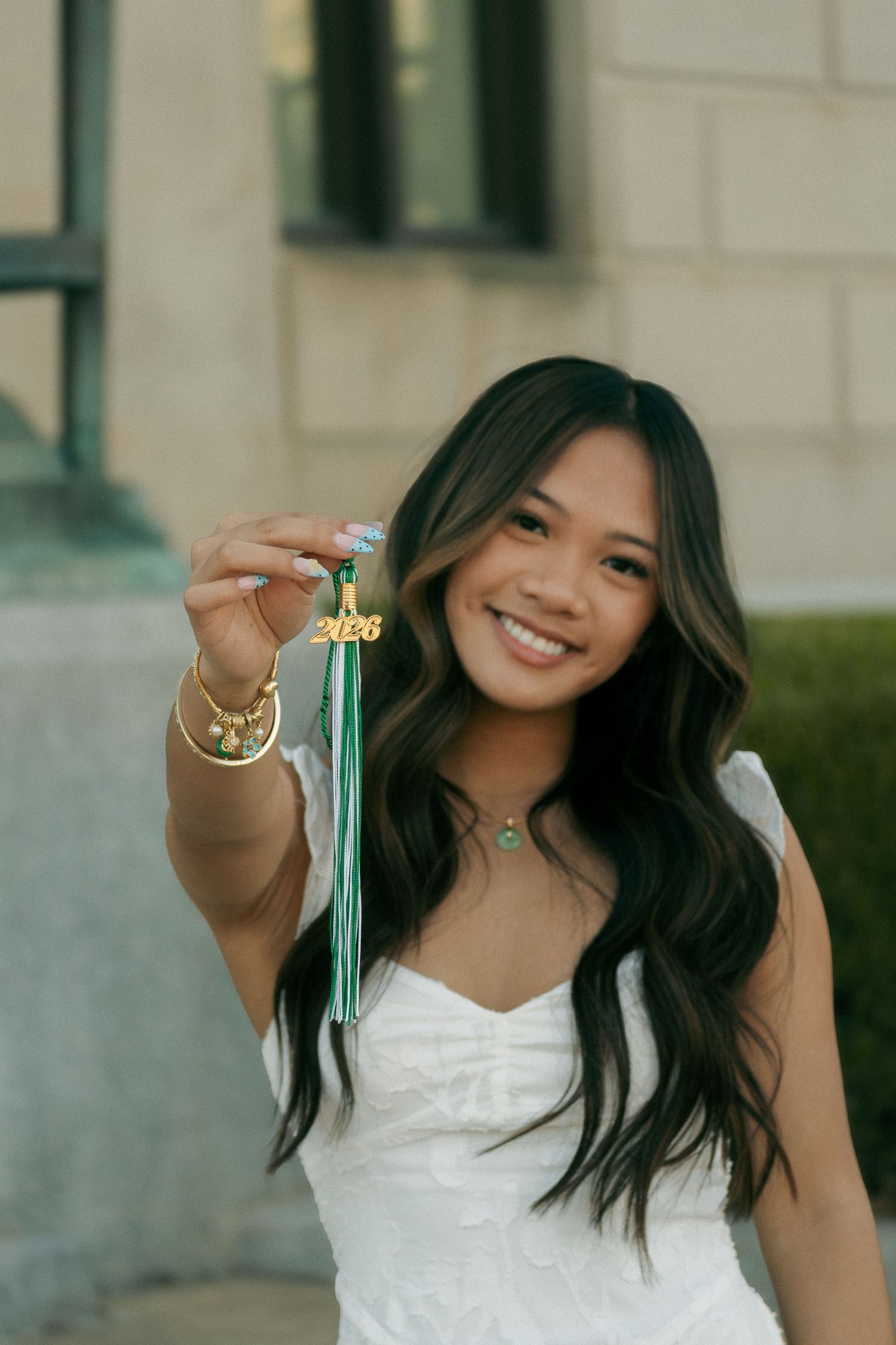 Smiling young woman in white dress holding green graduation tassel with 2026 charm for senior photos.