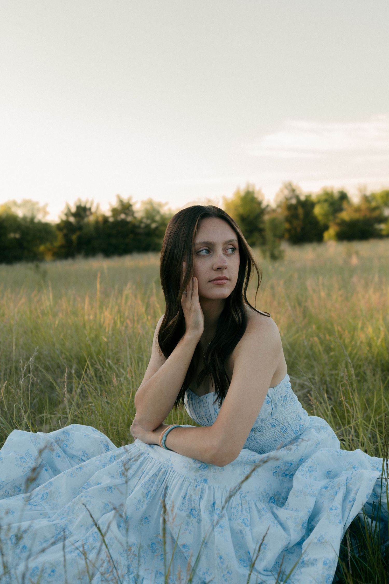 Young woman in a light blue floral dress sitting in a grassy field at dusk, gazing thoughtfully into the distance.
