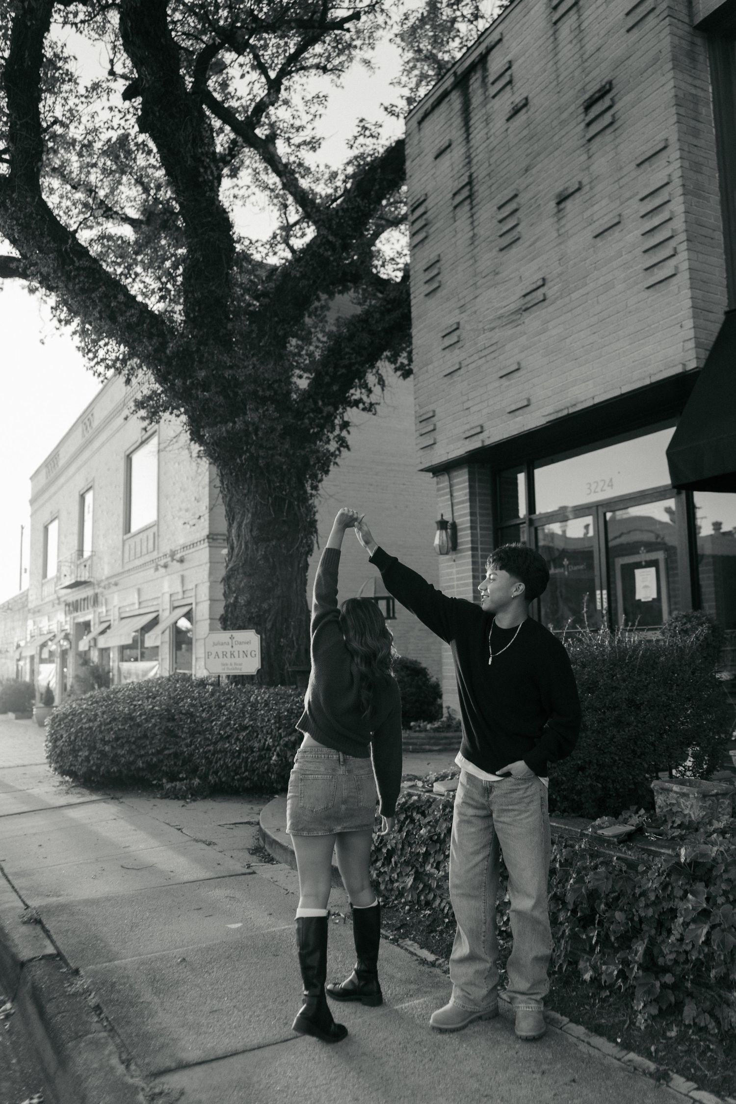 Black and white photo of a young man spinning his girlfriend on a city sidewalk near a large tree.