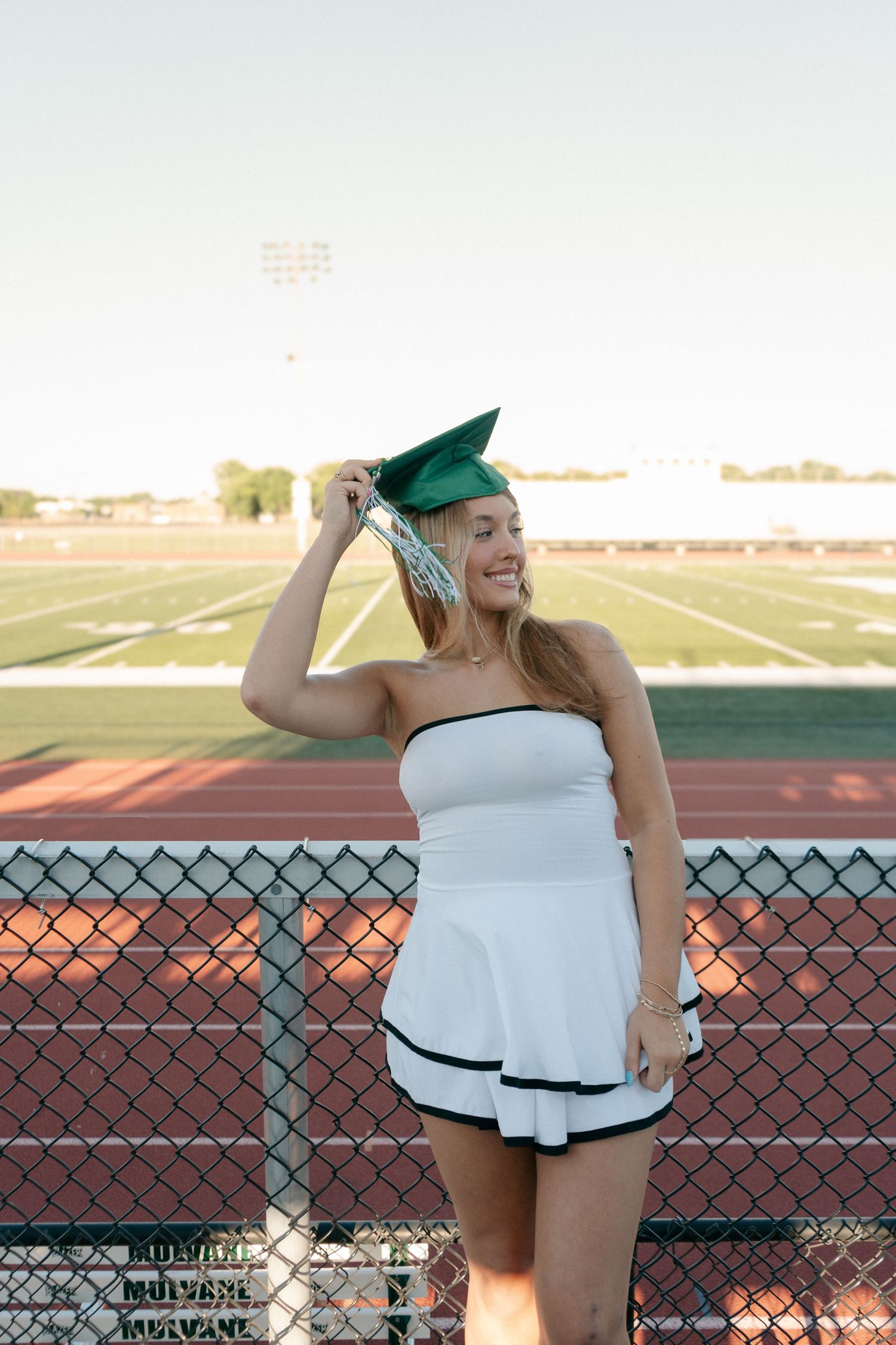 Smiling graduate in white dress holds green cap at stadium track.