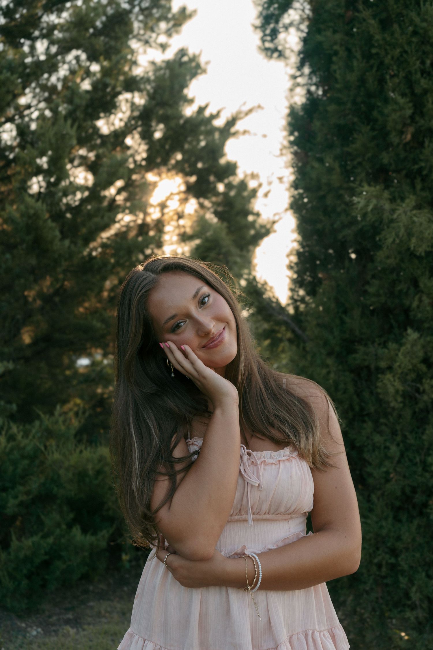 Smiling young woman in blush pink dress resting hand on cheek posing outdoors at golden hour sunset.