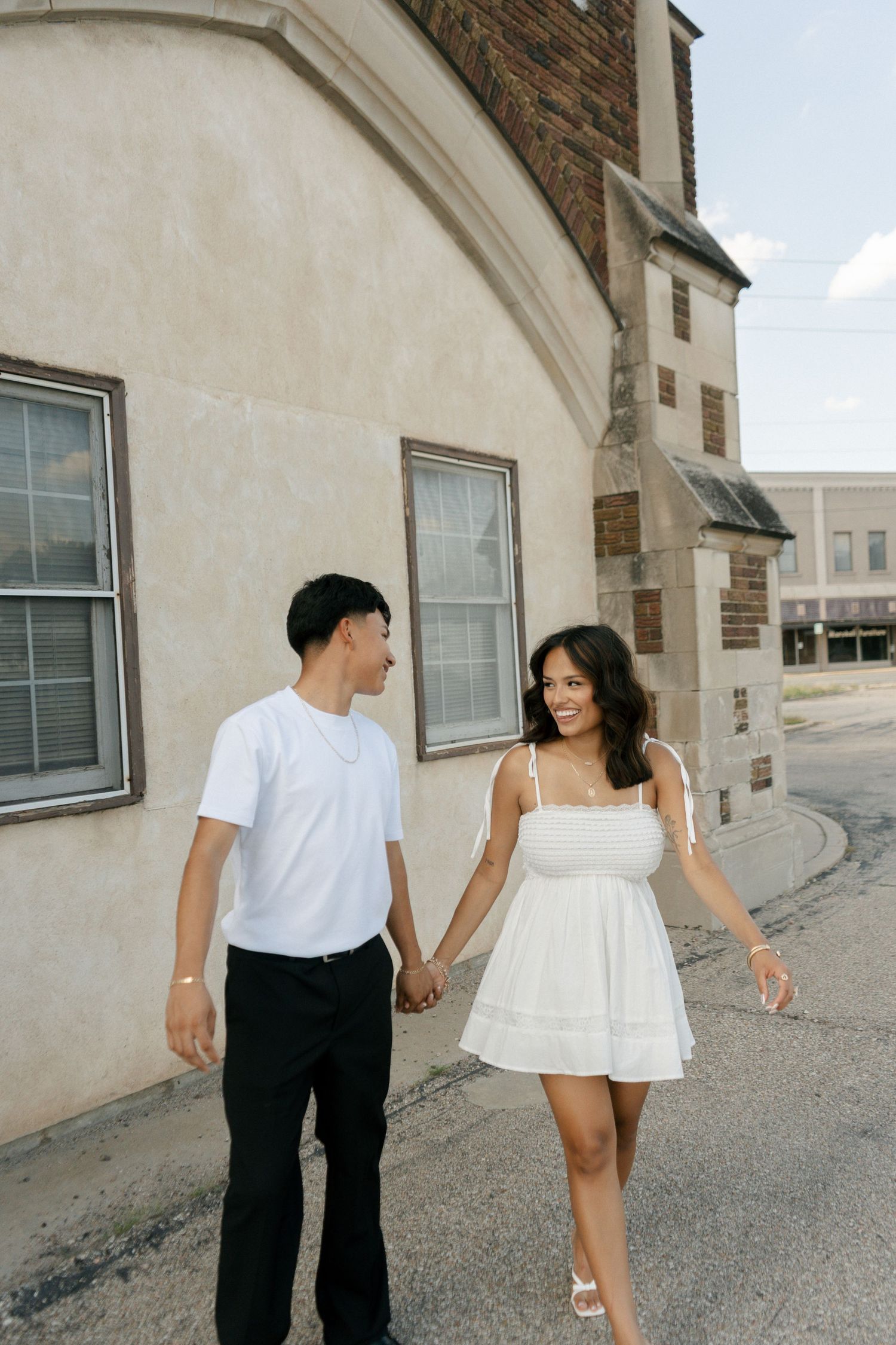 Couple holding hands and smiling at each other while walking along a street beside a beige building.