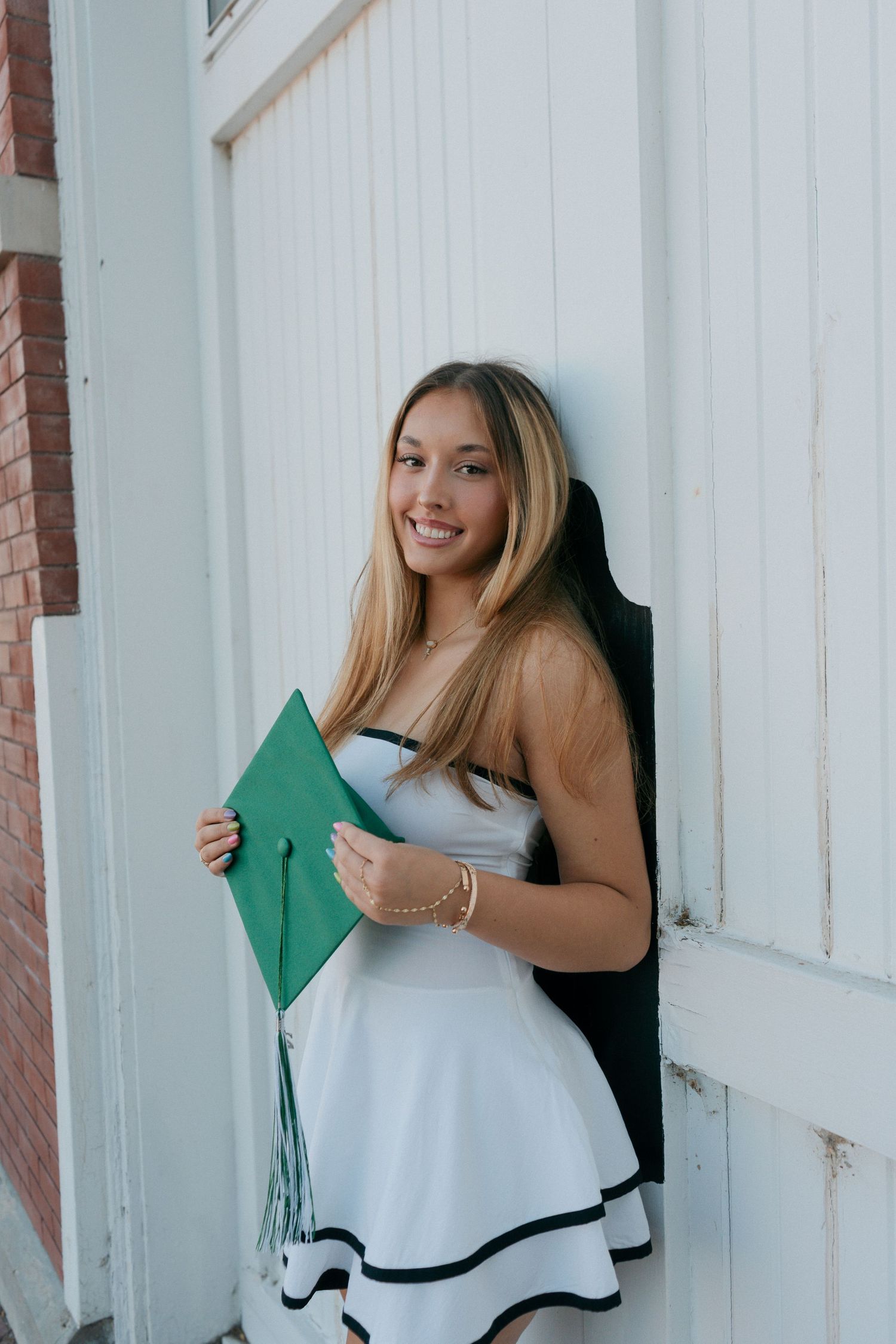 Happy graduate in white dress leaning against white door holding green mortarboard.