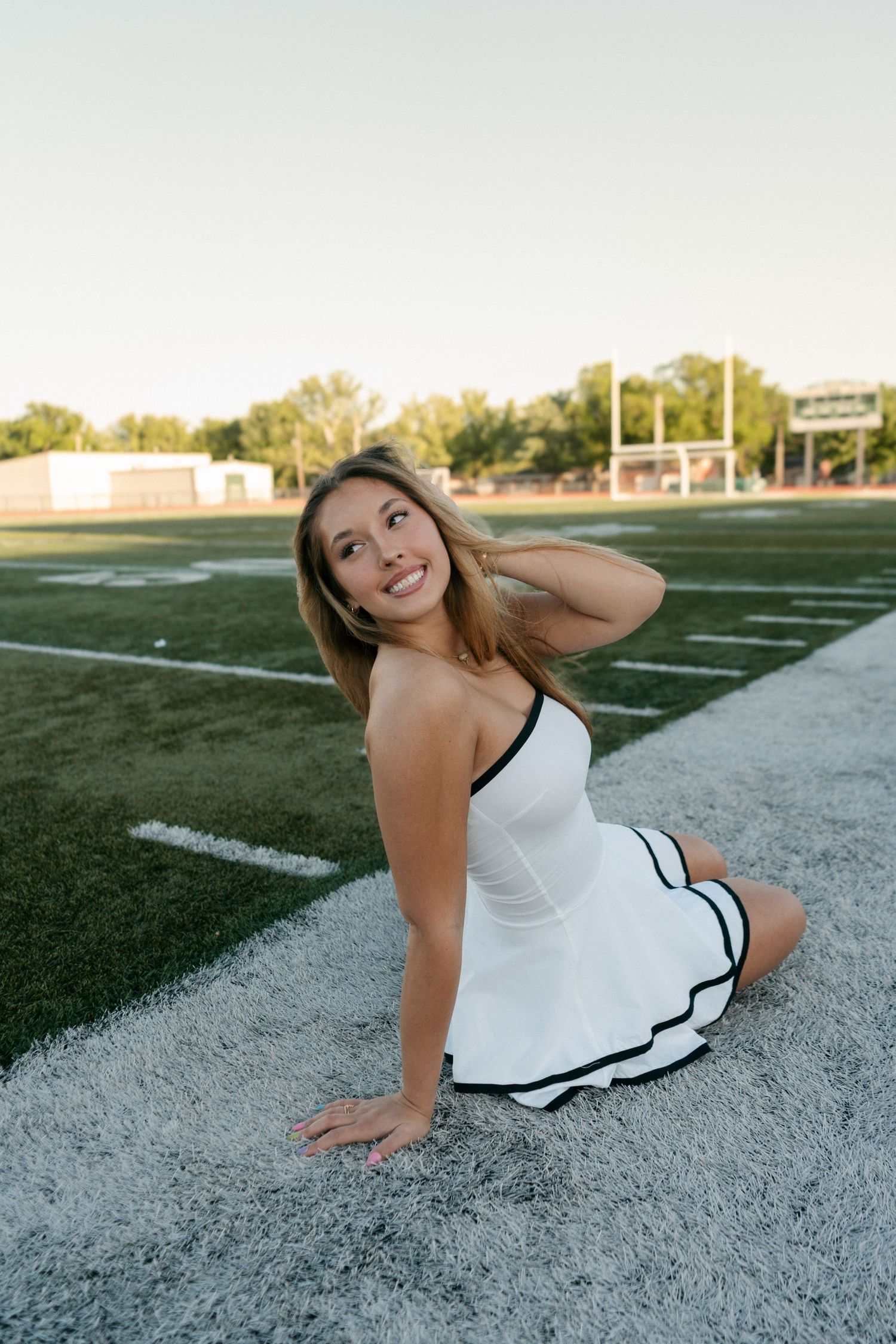 Smiling young woman in white dress posing on green football field turf.