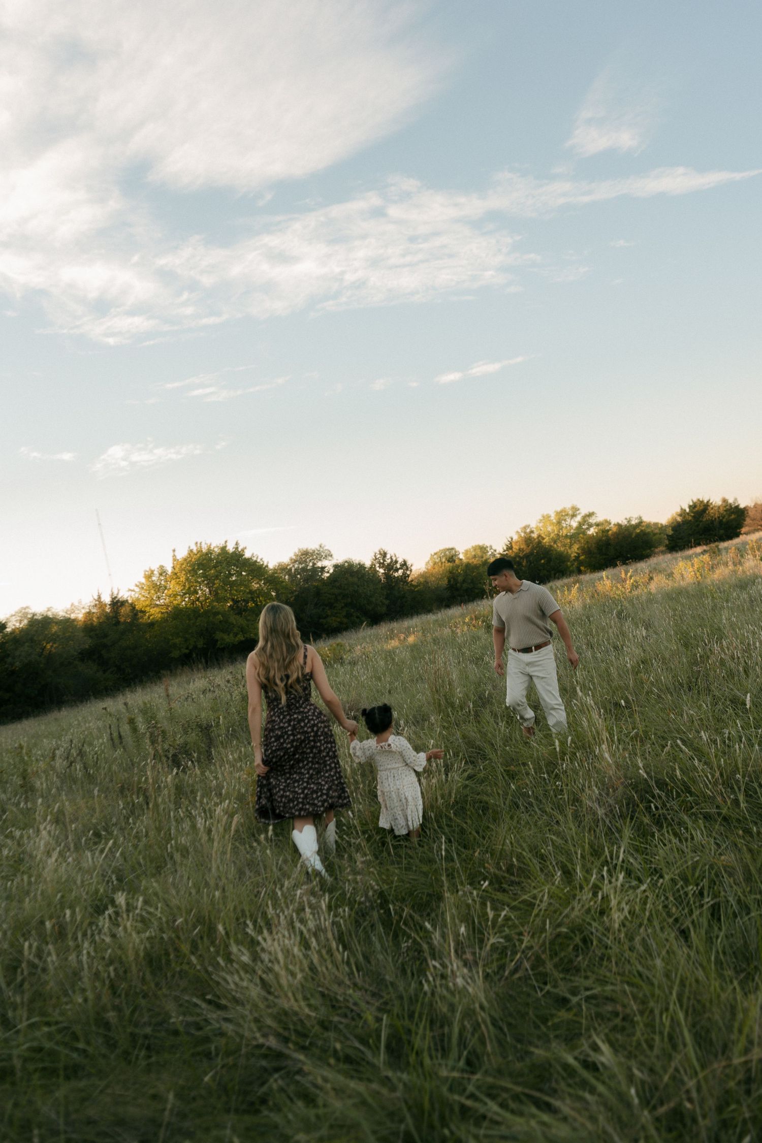 A family of three walks hand in hand through a wide open green meadow under a partly cloudy sky.