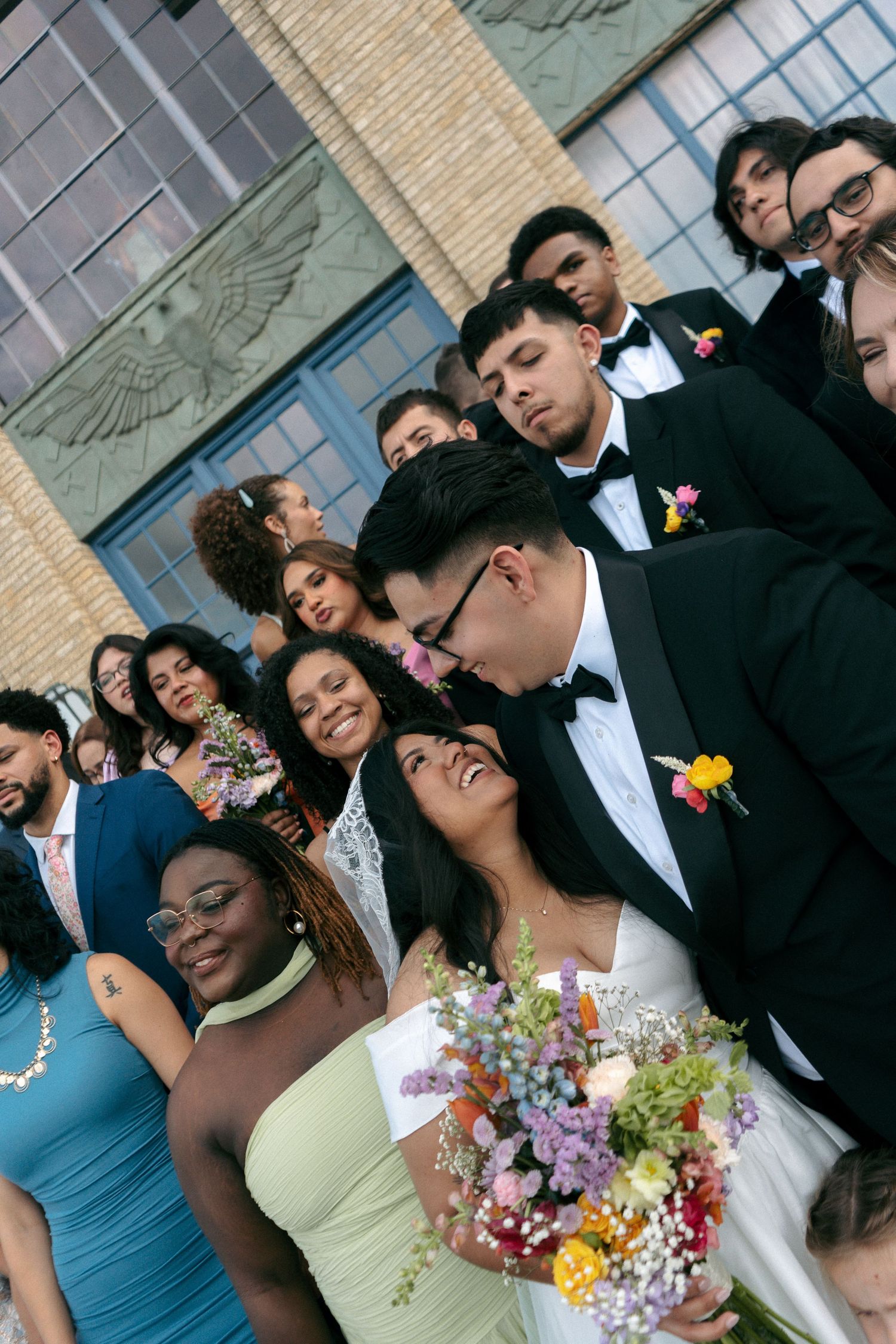 Couple shares a kiss surrounded by smiling wedding guests outside a brick building, bride holding colorful bouquet.