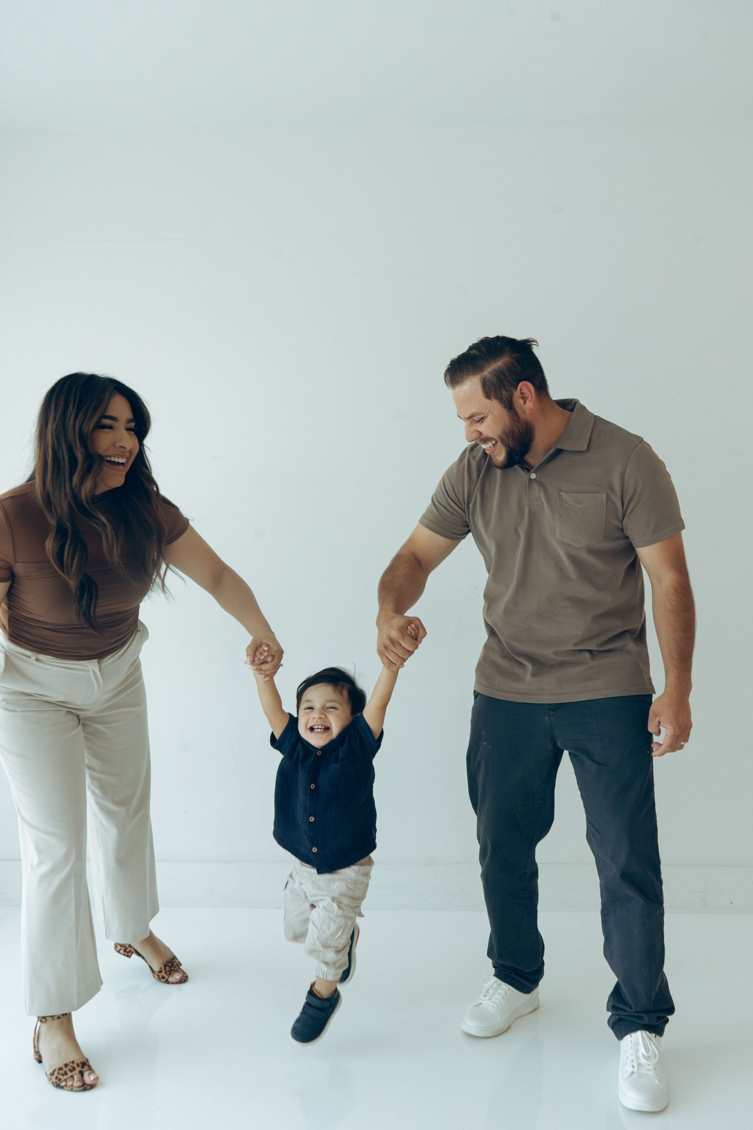 Happy family with parents swinging toddler boy by hands in bright white studio.