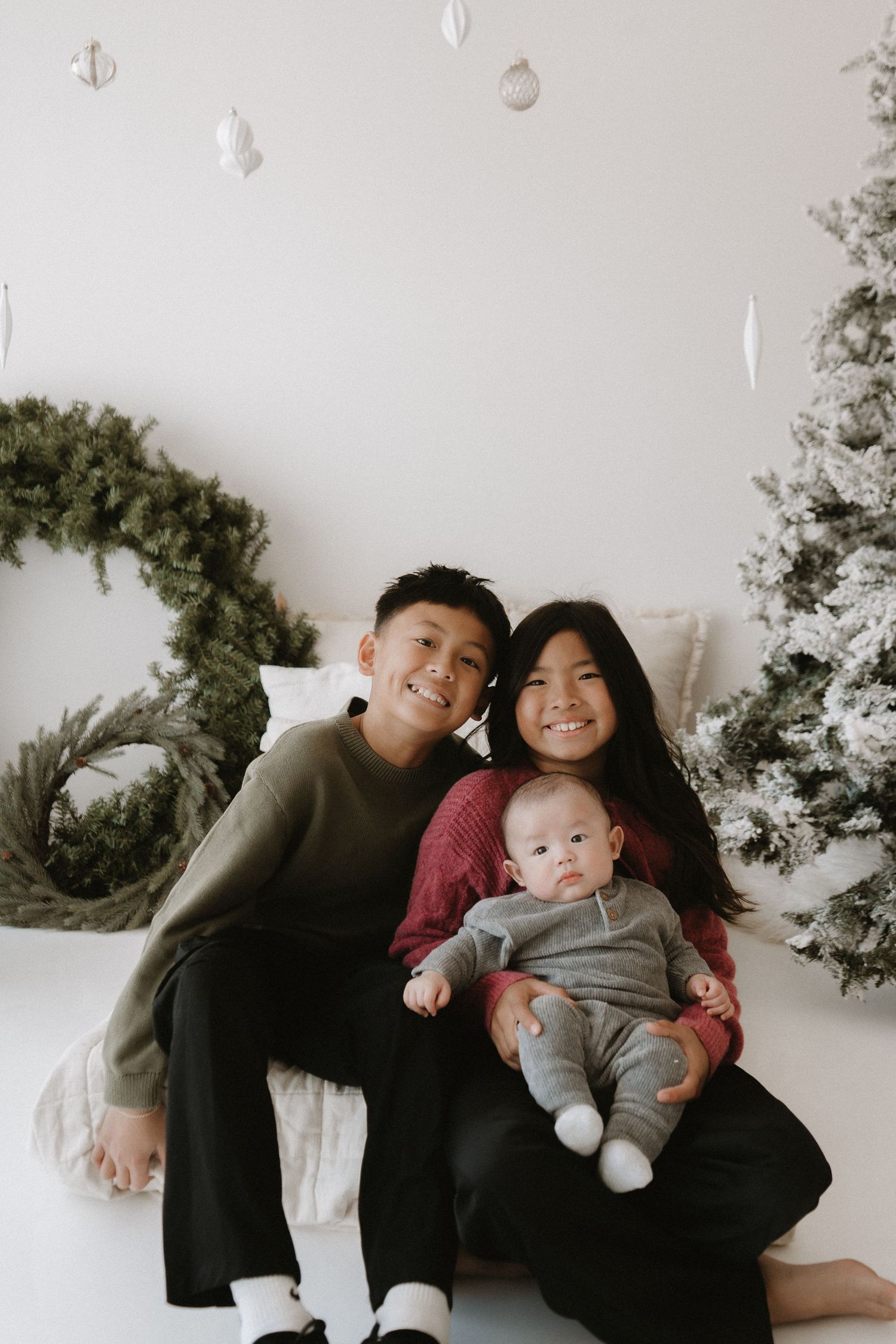 Two Asian children sit together holding an infant sibling in front of a snow-flocked Christmas tree.