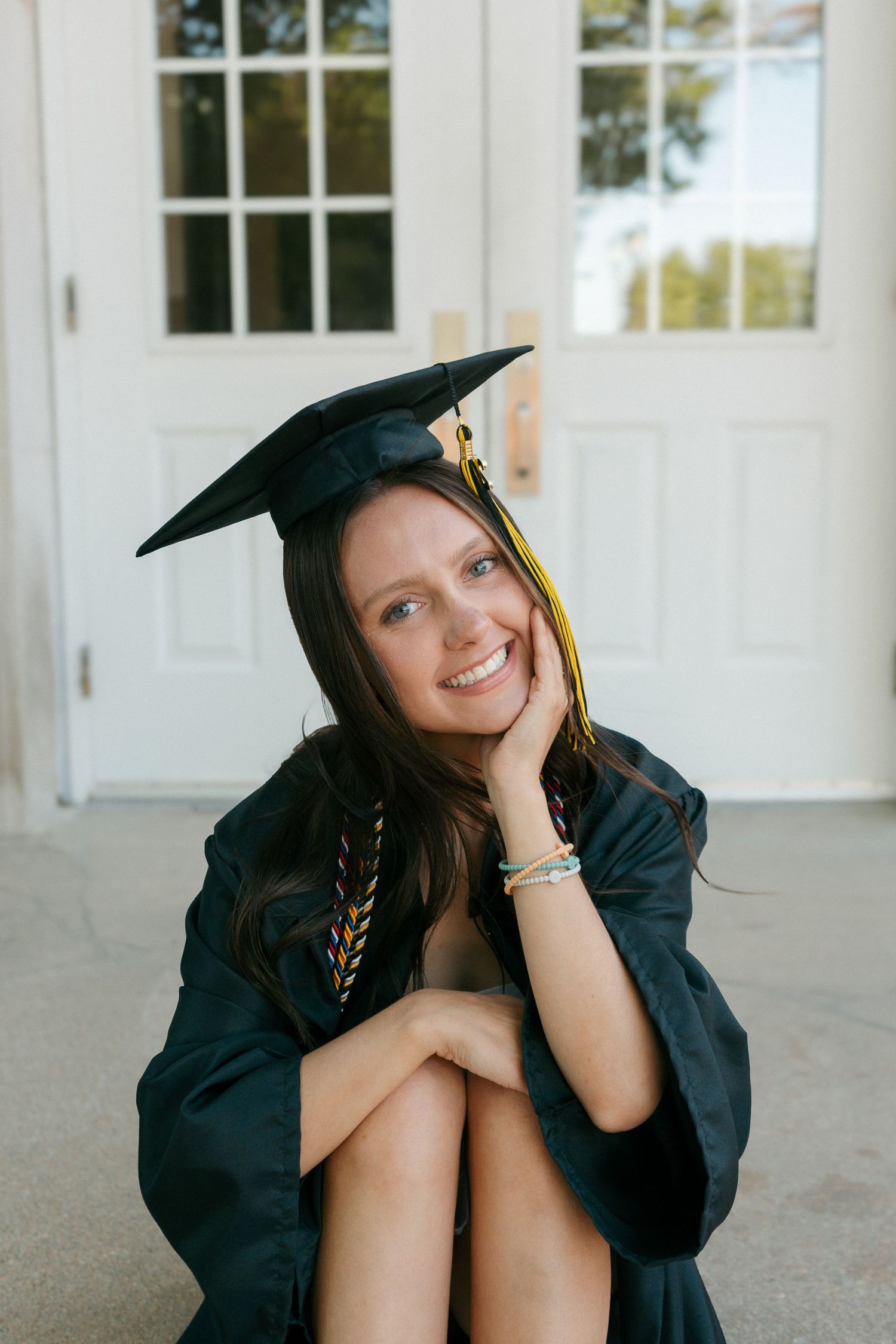 Smiling young woman in black graduation cap and gown sitting outdoors, celebrating her academic achievement.