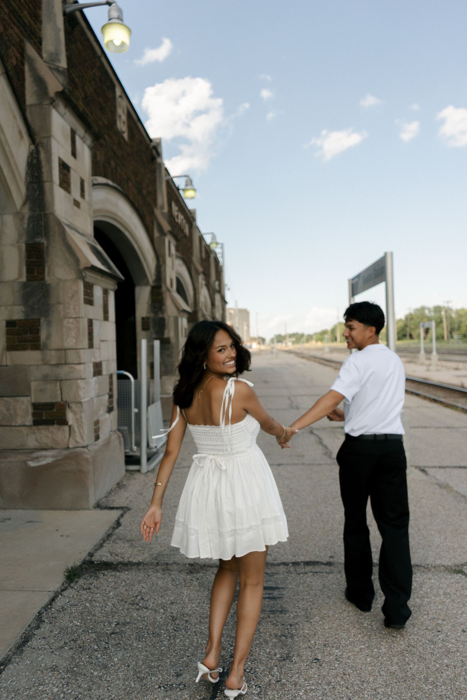 Couple holding hands at a train station, woman in white dress smiling back at her partner.