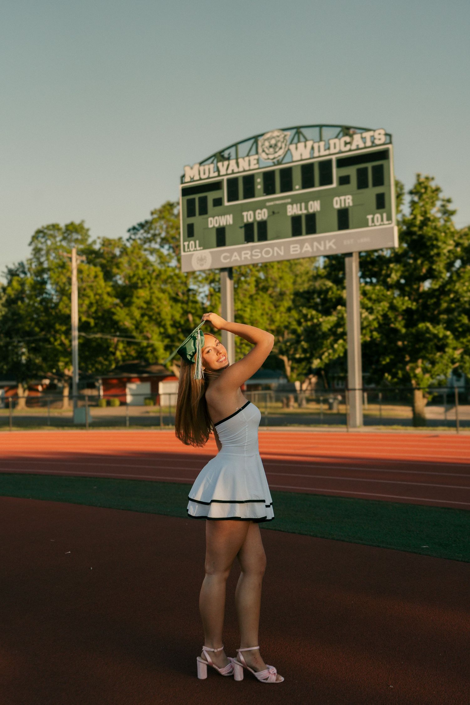 Young woman in white dress posing near Mulvane Wildcats scoreboard at golden hour.