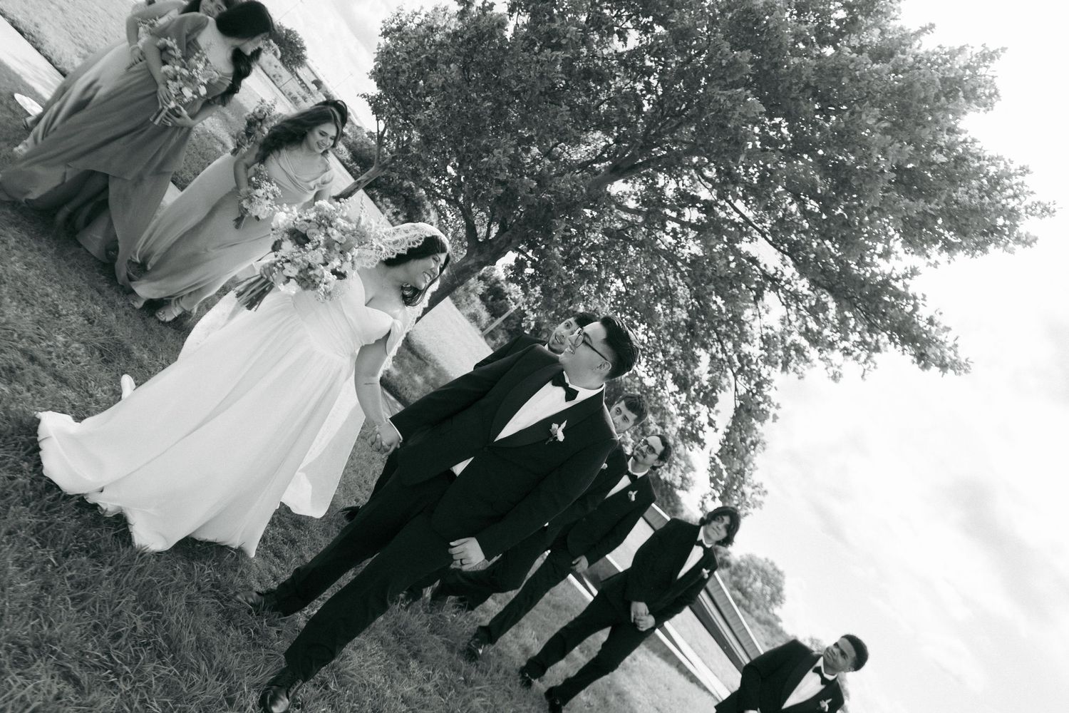 Black and white wedding party photo outdoors near a tree, featuring bride, groom, bridesmaids, and groomsmen.