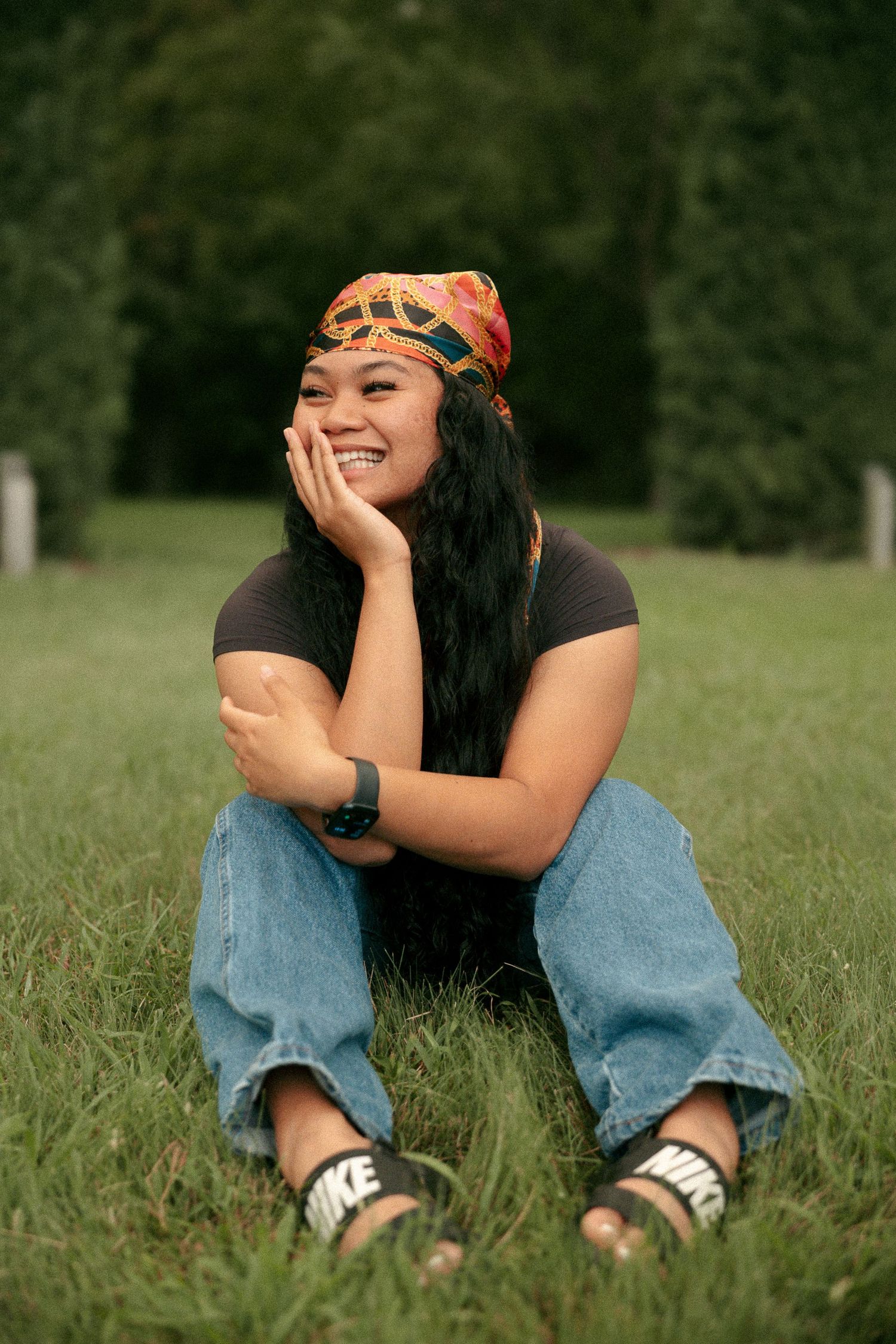 Smiling young woman sitting on grass wearing colorful headband, black tee, jeans, and Nike sandals.