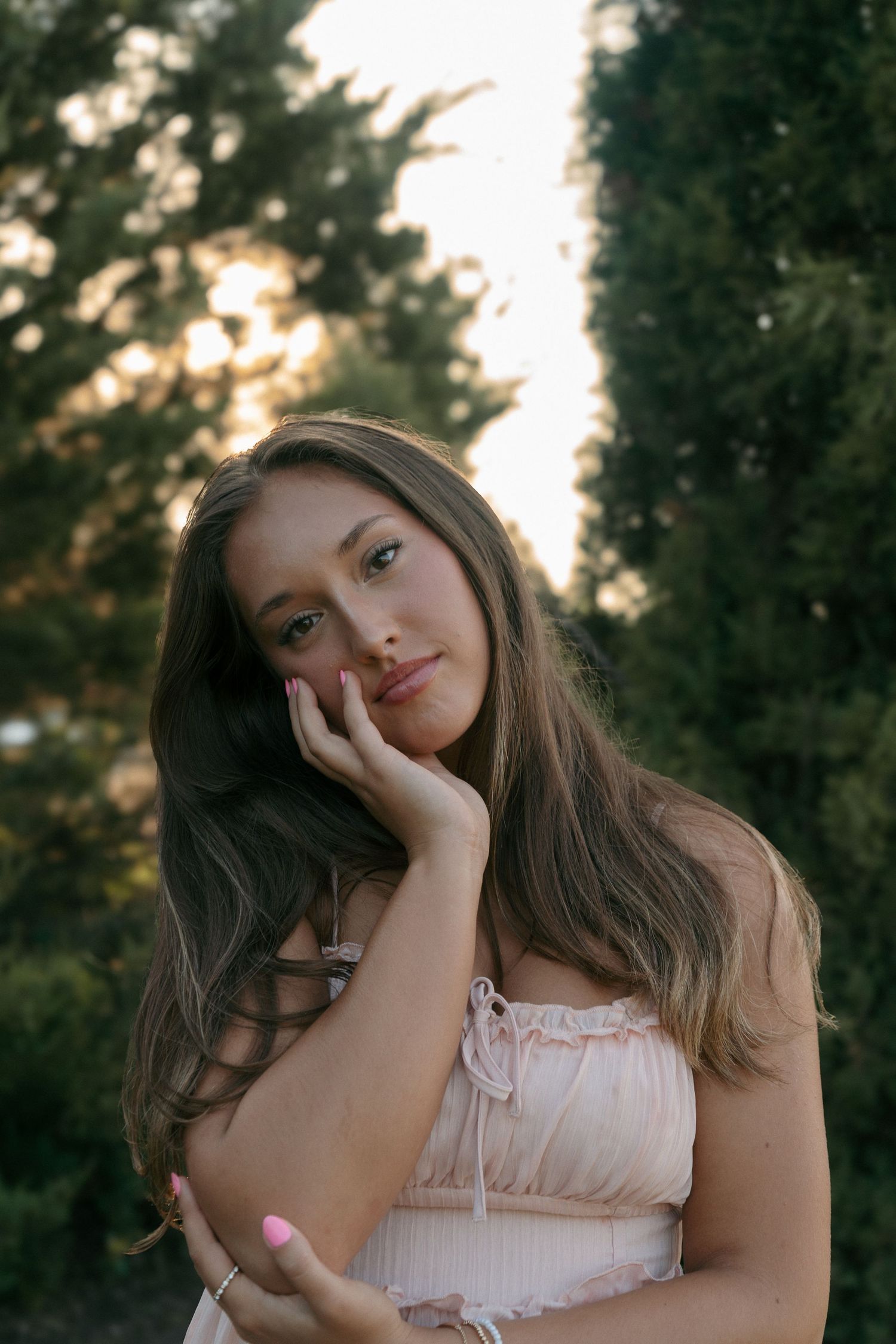 Young woman in pink ruffled dress resting hand on cheek posing outdoors among evergreen trees at sunset.