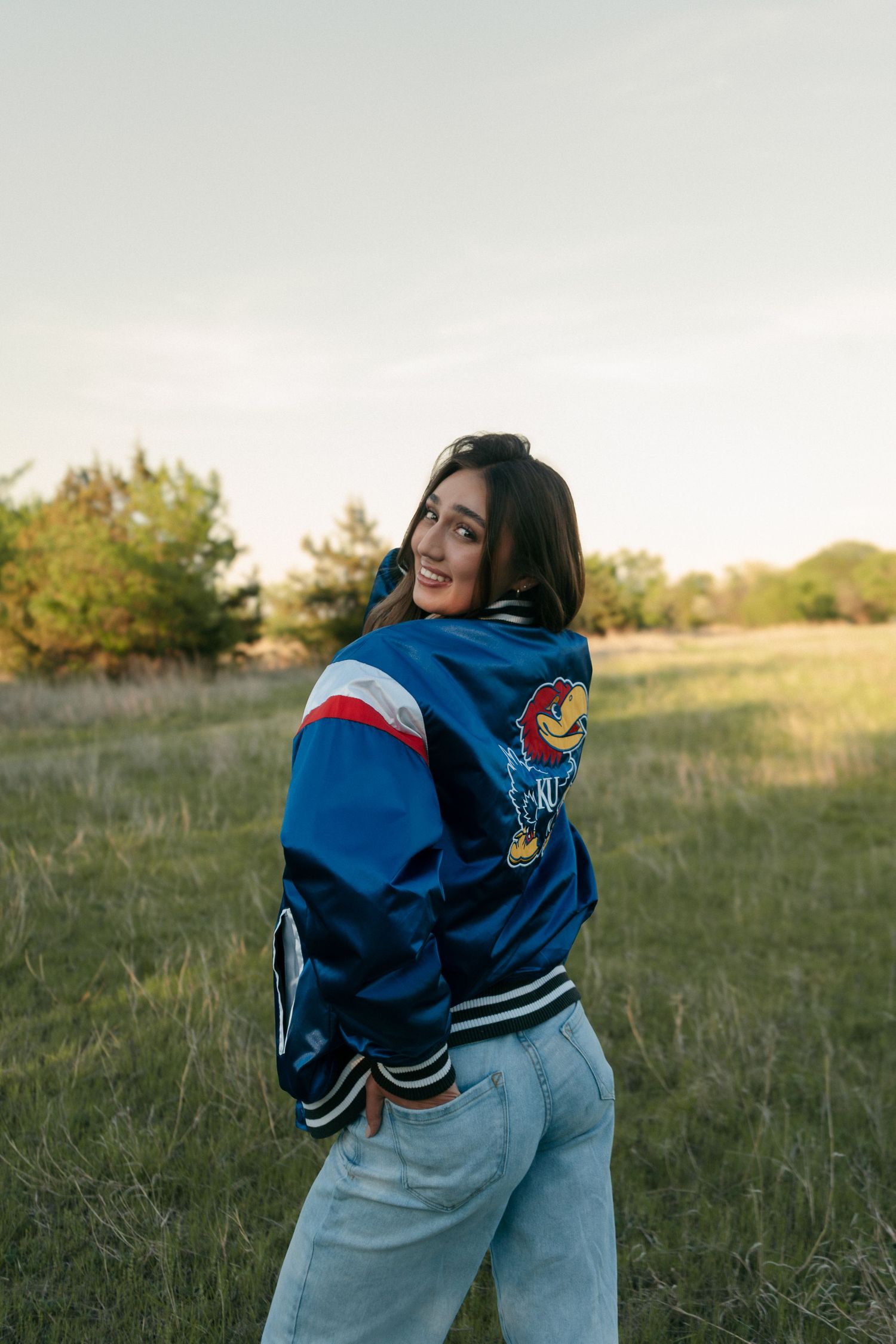 Young woman smiling over shoulder in blue Kansas Jayhawks varsity jacket in a green field at sunset.