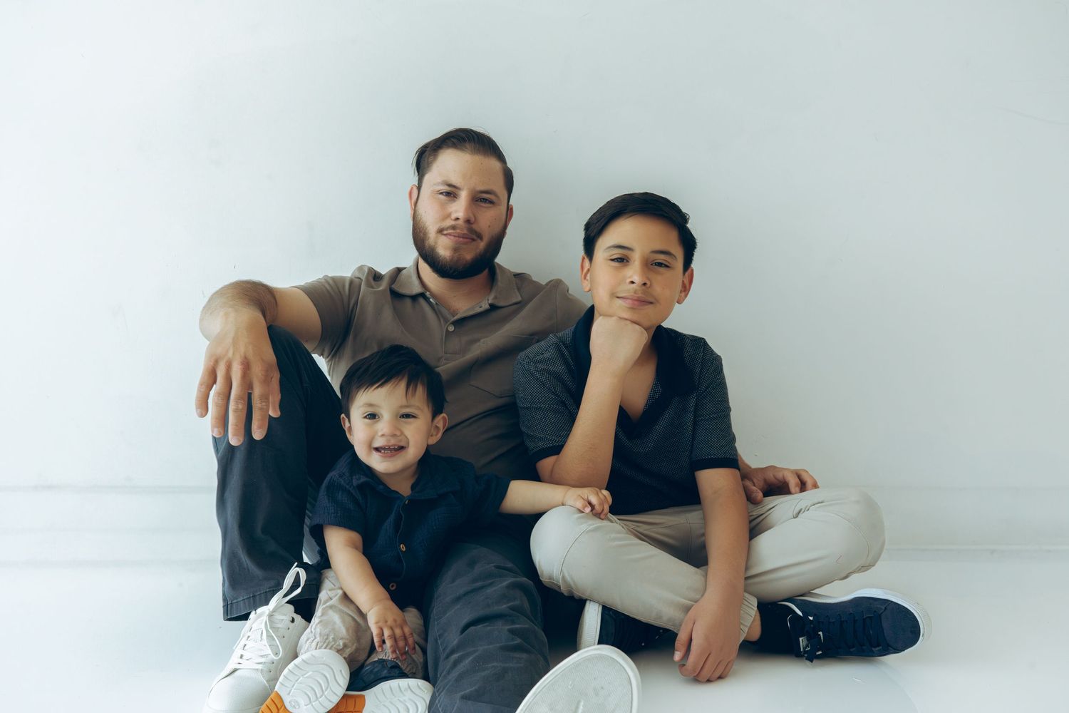 Father sitting on floor with two young sons posing for portrait in white studio.