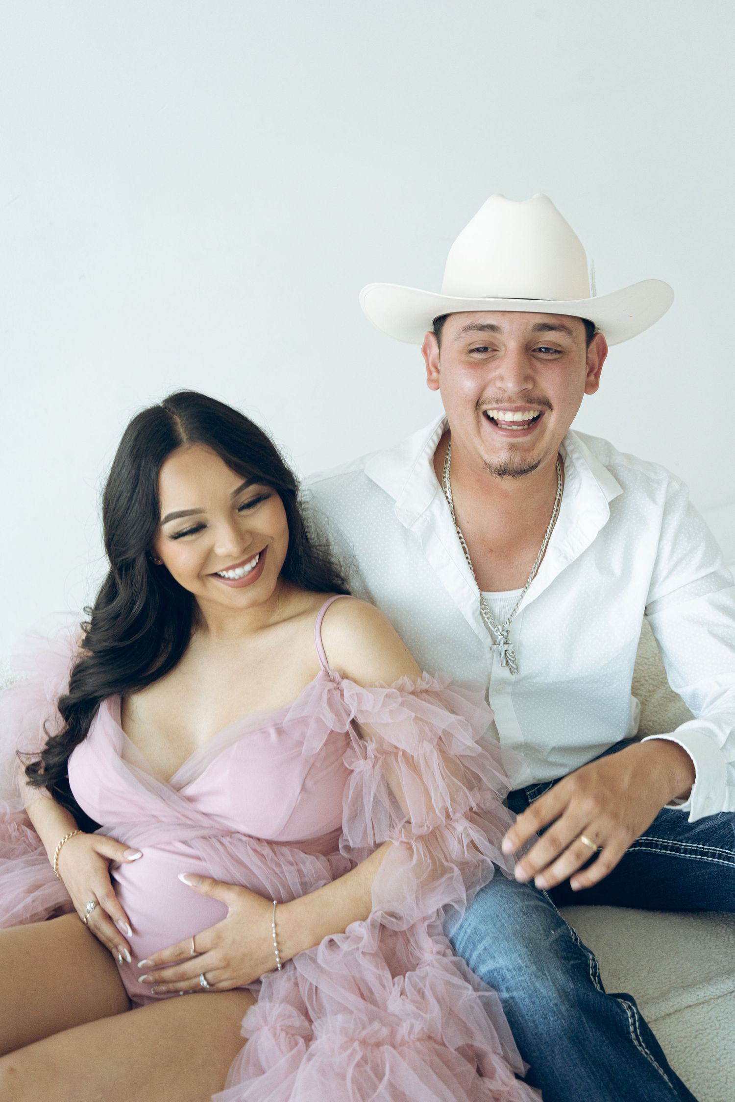 Laughing expectant couple in pink tulle dress and white cowboy hat on couch.