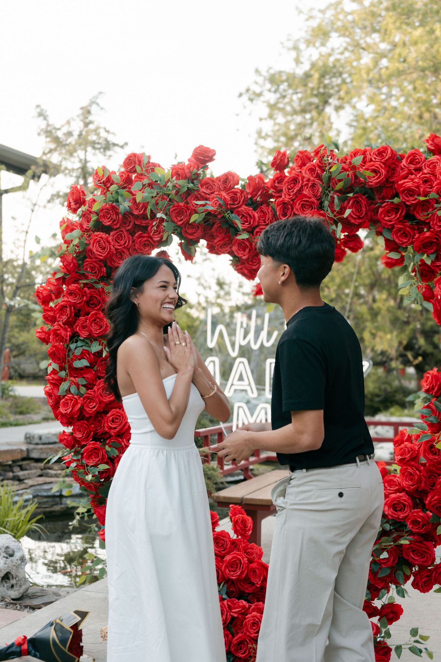 Emotional woman clasping hands as man prepares to propose under stunning red rose heart-shaped arch.
