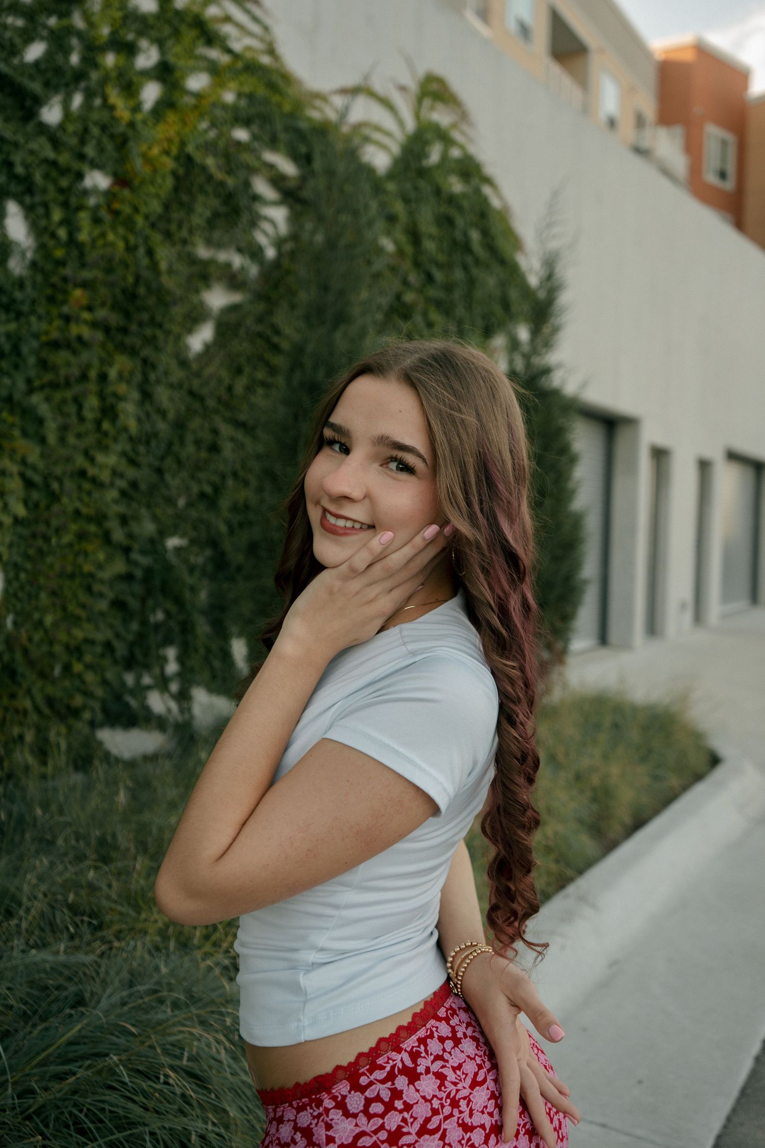 Young woman in white crop top and red floral skirt smiling beside an ivy-covered building.