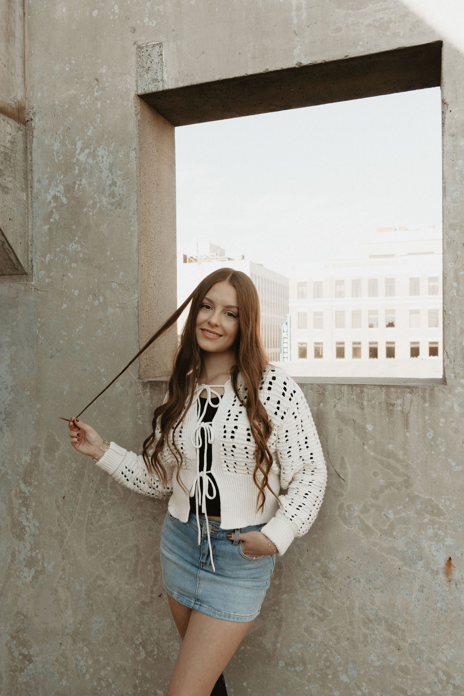 A young woman in a white crochet top and denim skirt poses against a concrete wall on a rooftop.